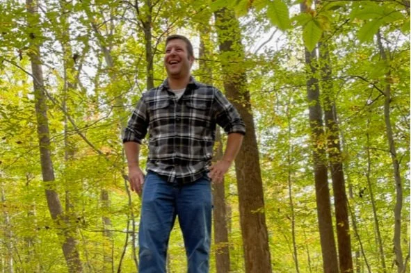 Matthew Peek enjoying an early fall hike. Matthew is standing on a fallen tree trunk, striking a confident pose, and laughing.