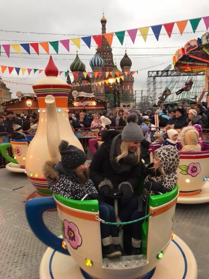 Crowded winter amusement park with a mother and two children riding a colorful teacup ride in front of Saint Basil’s Cathedral in Moscow