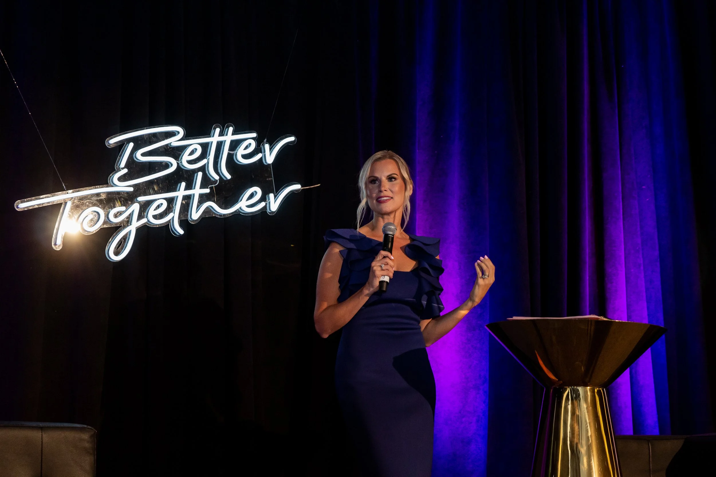 Blonde woman in a navy ruffled dress speaking into a microphone onstage beside a lit “Zetter Lighter” sign and gold cocktail table.