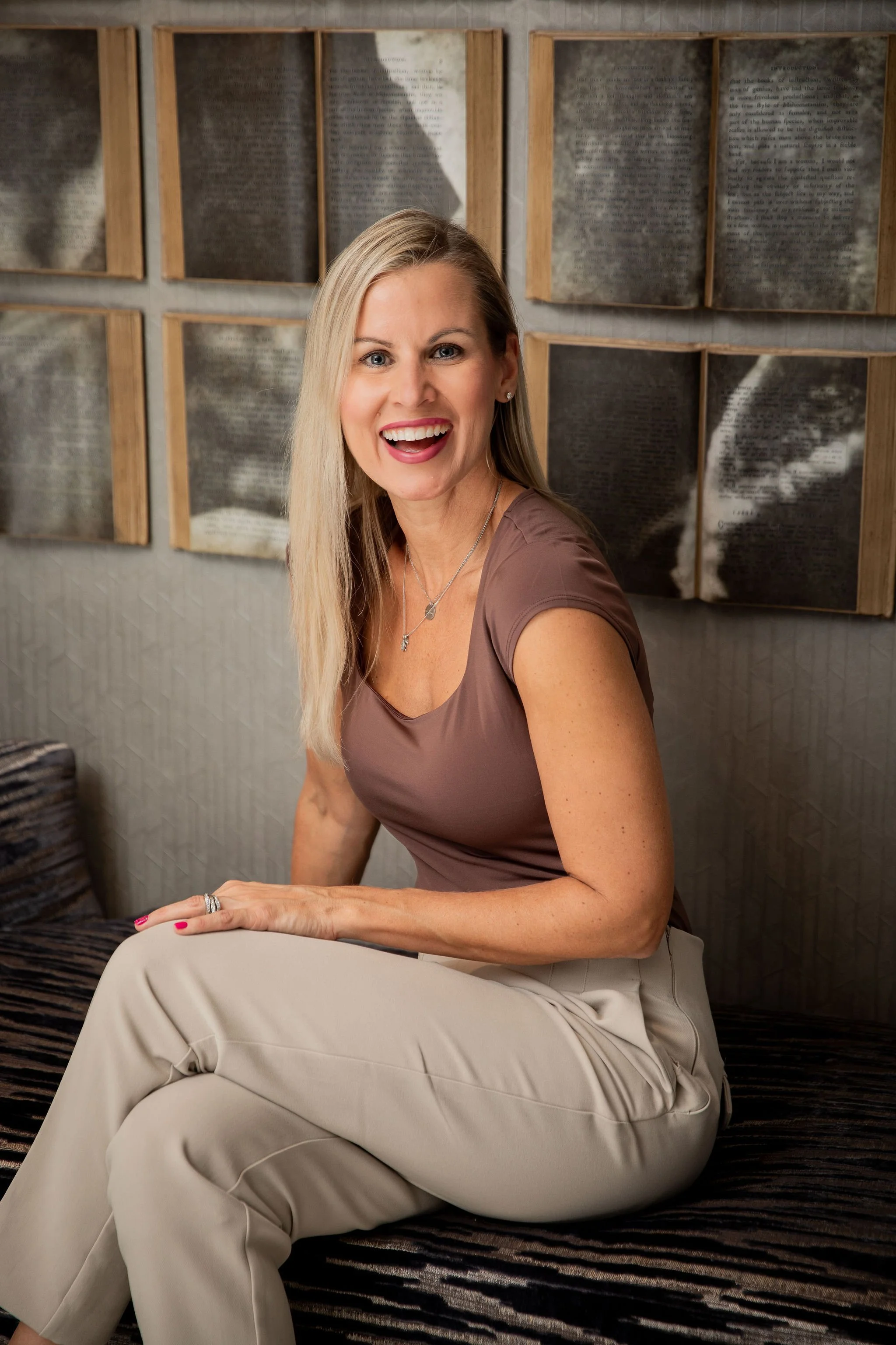 Smiling blonde woman seated indoors in front of framed book pages.