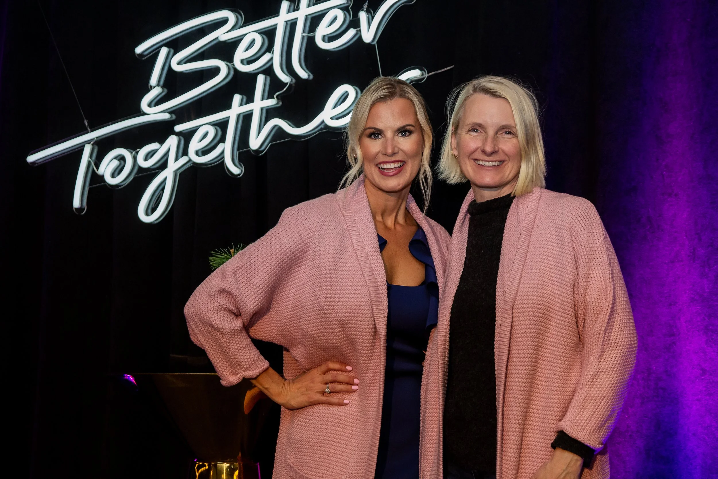 Two smiling women in pink cardigans pose beneath a glowing “Better Together” sign at an event.