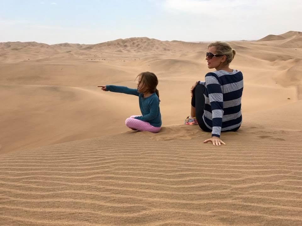 Mother and daughter sitting on a rippled sand dune in a desert, with the child pointing toward the landscape.