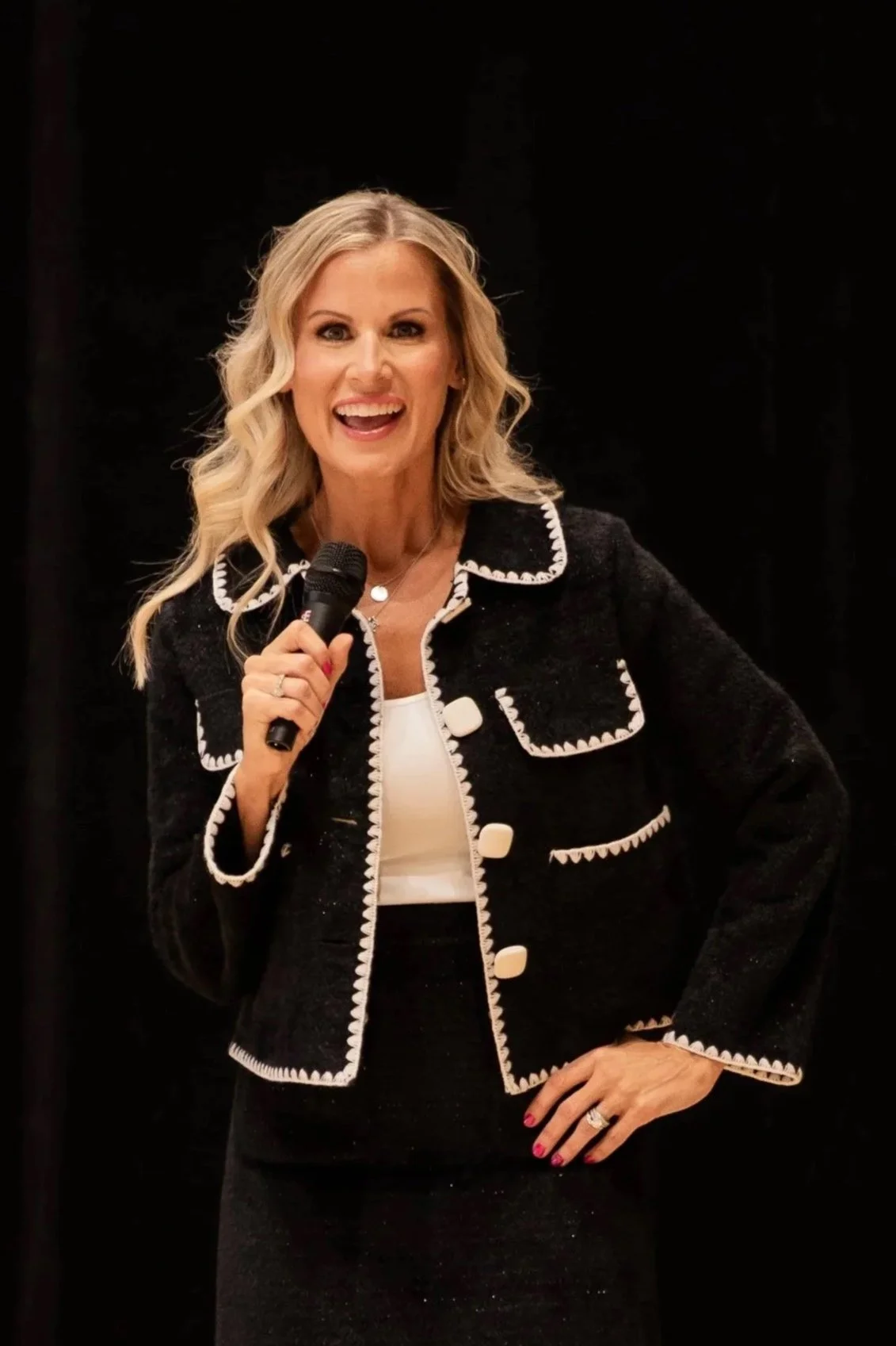 Blonde woman smiling and speaking into a microphone while wearing a black jacket with white trim on a dark stage