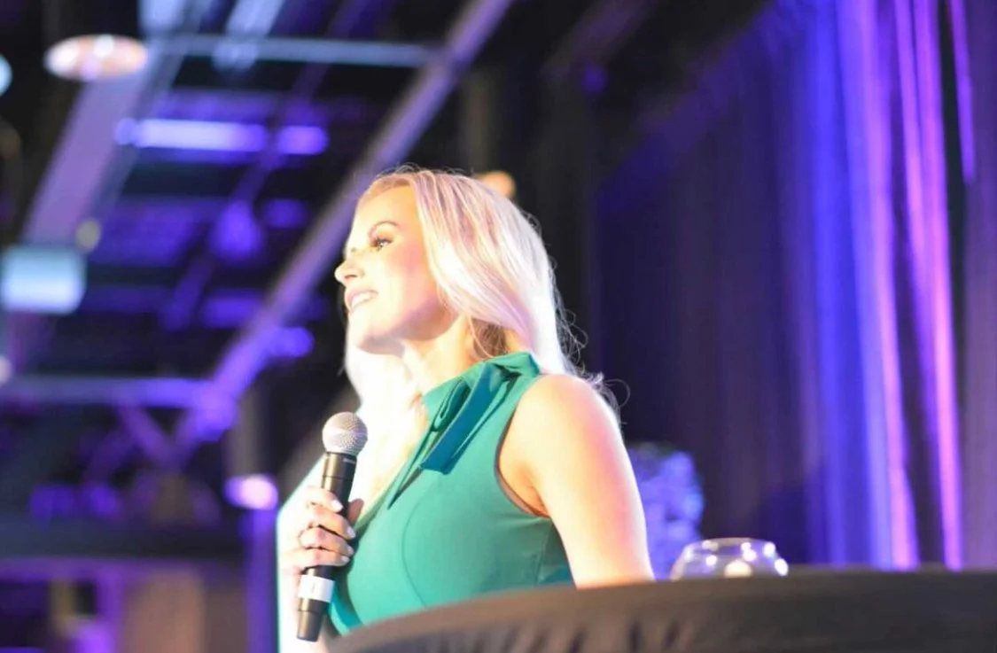 Blonde woman in a teal dress singing into a microphone on a purple-lit stage