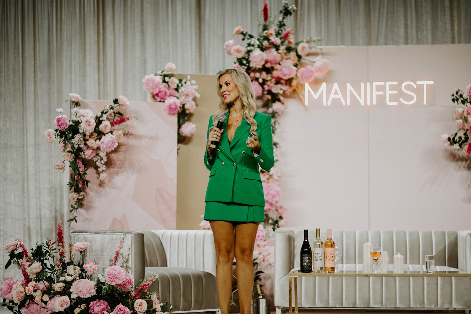 Woman in a green suit speaking into a microphone on a flower-decorated stage labeled “MANIFEST,” with sparkling wine bottles and glasses nearby