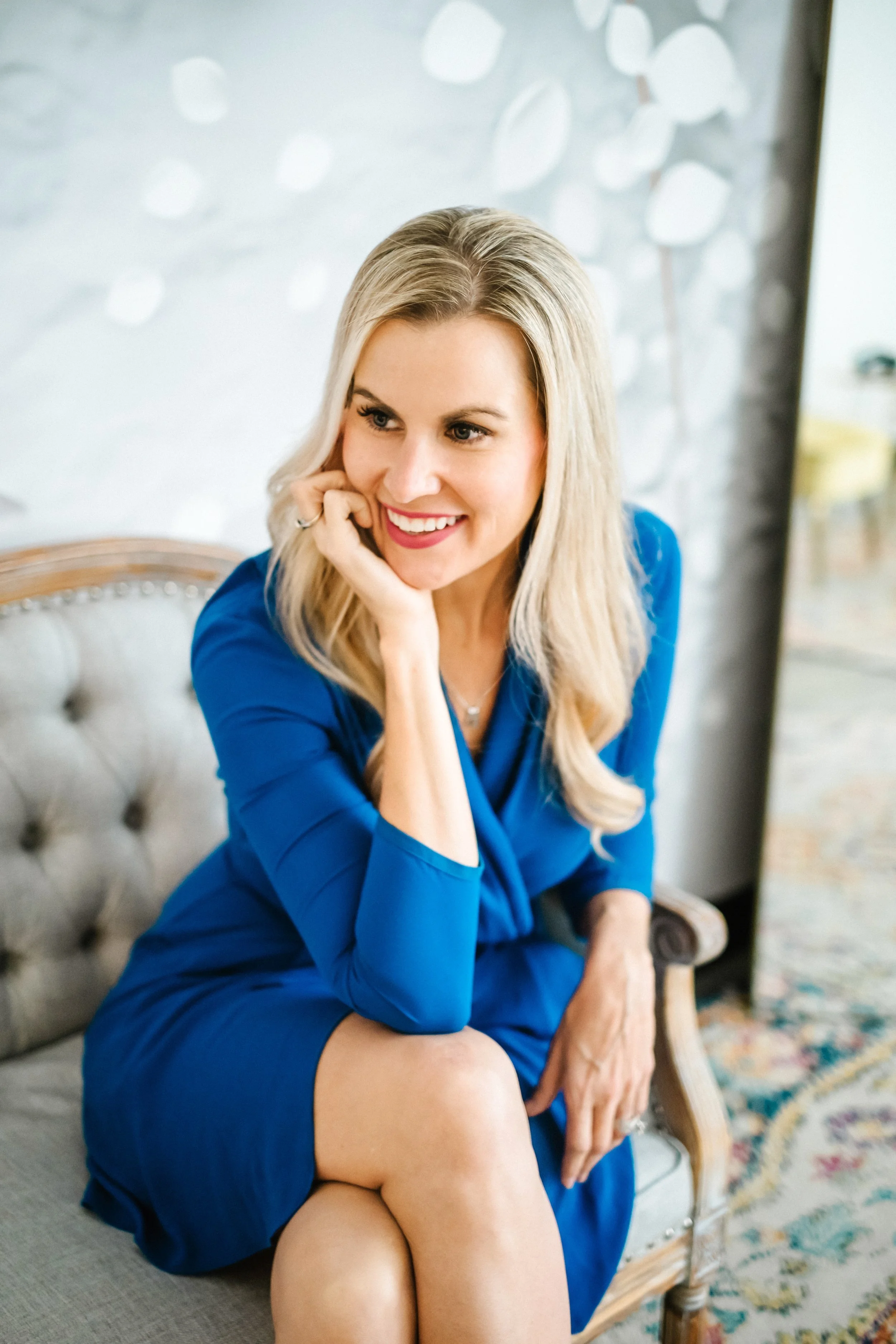 Smiling blonde woman in a blue dress sitting indoors and resting her chin on her hand.
