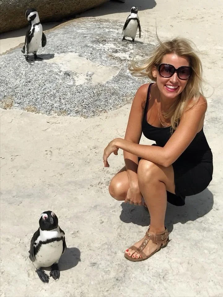 Lisa Webb, keynote speaker and master of ceremonies in sunglasses crouching beside three African penguins on a sandy beach