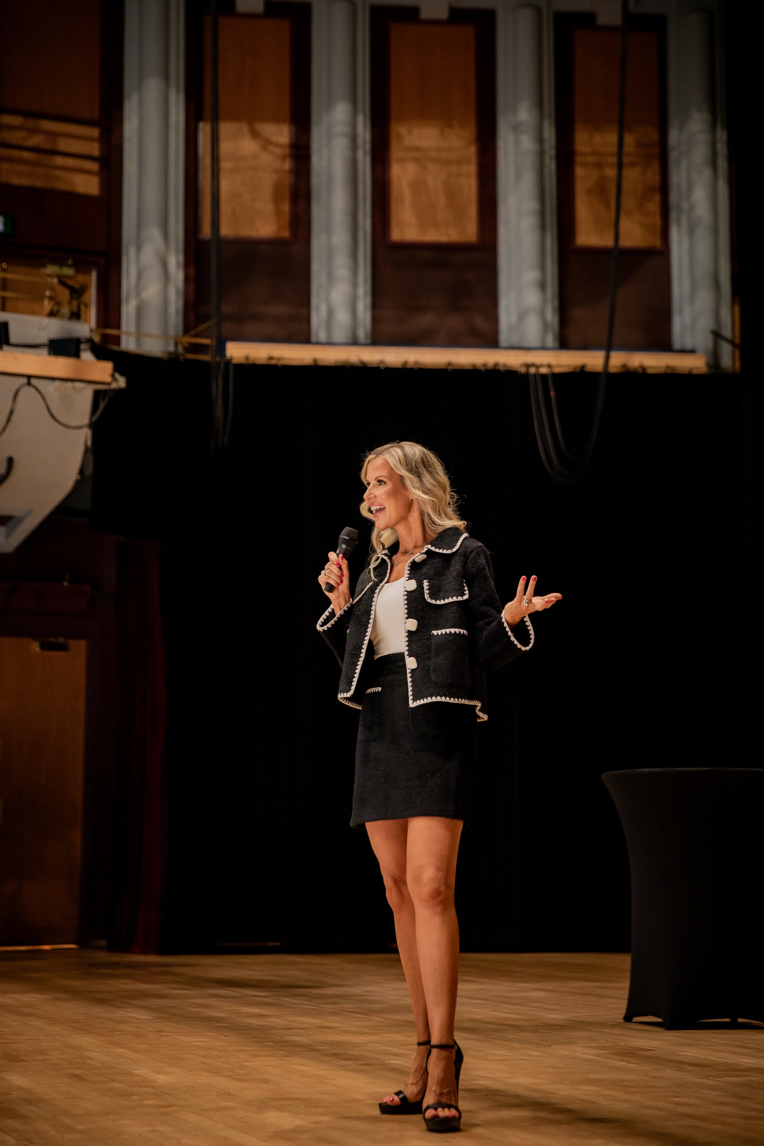 Blonde woman speaking into a microphone on a theater stage, wearing a black-and-white outfit.
