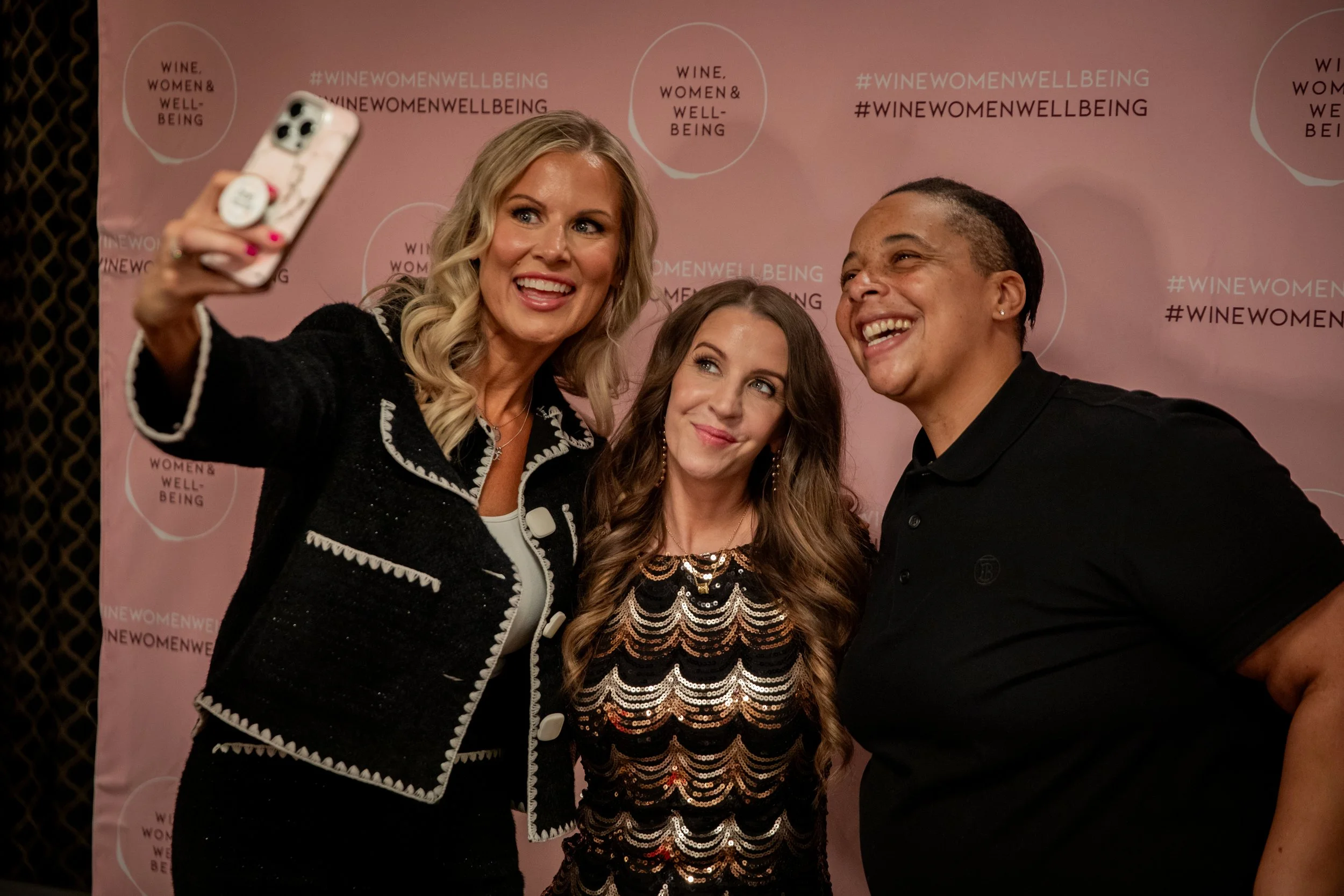 Four people take a selfie at a Wine Women & Wellbeing event in front of a pink branded backdrop.