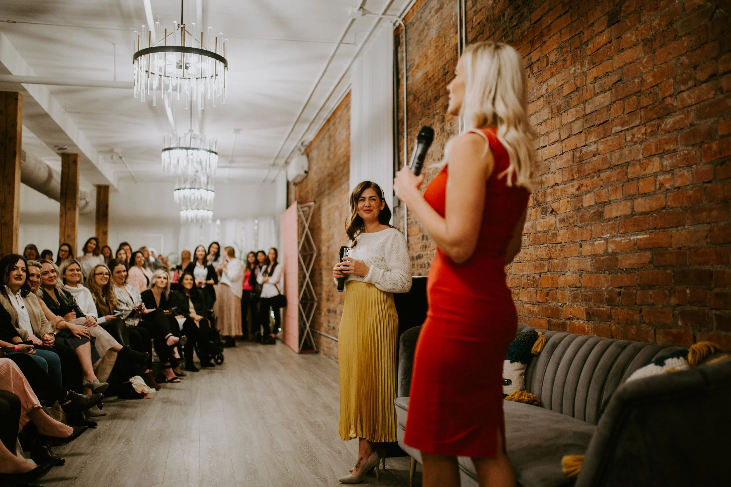 Woman in a red dress speaking into a microphone at a fashion event while an audience watches in an industrial brick-walled venue.