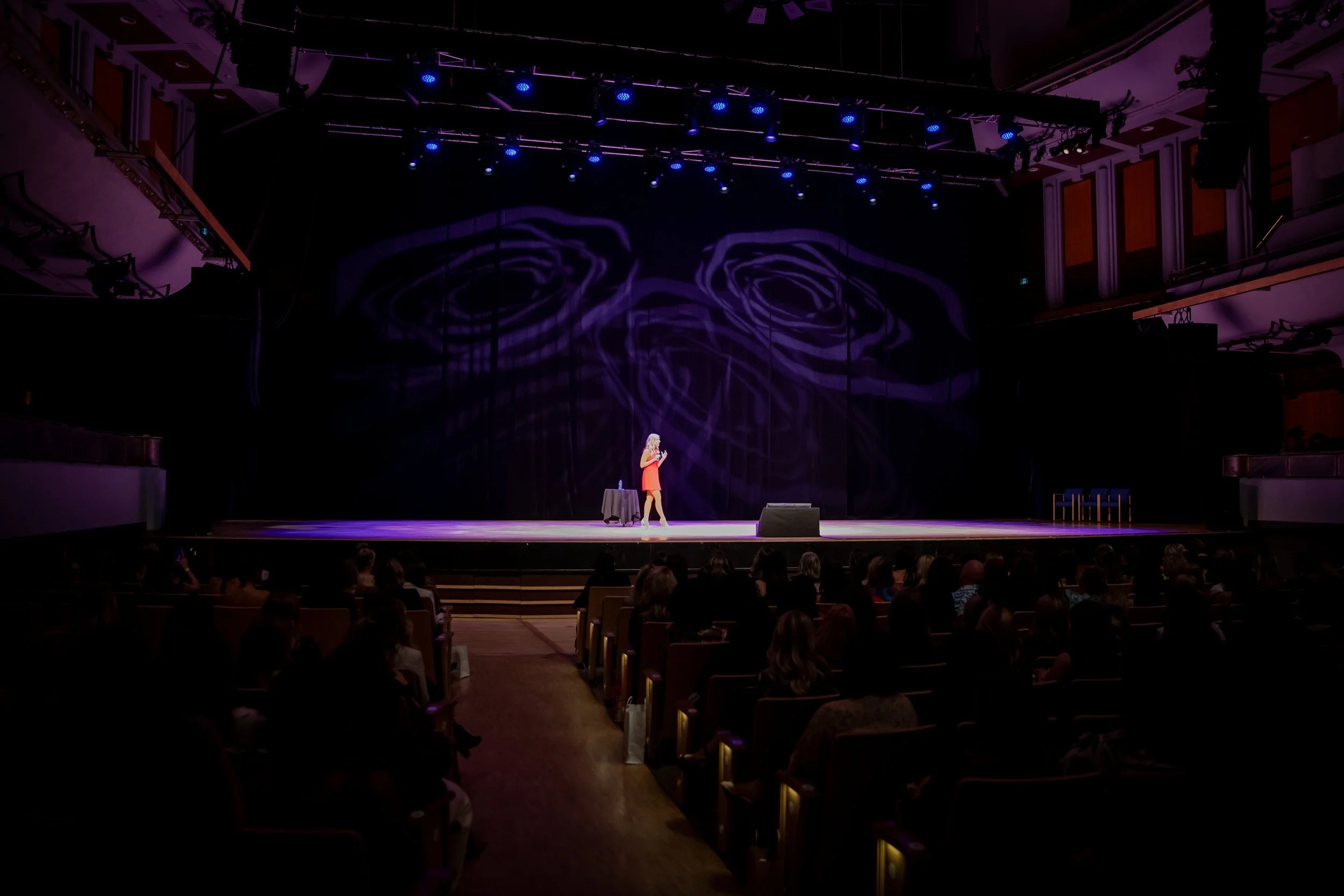 Woman speaking on a theater stage before a seated audience under purple and blue lighting