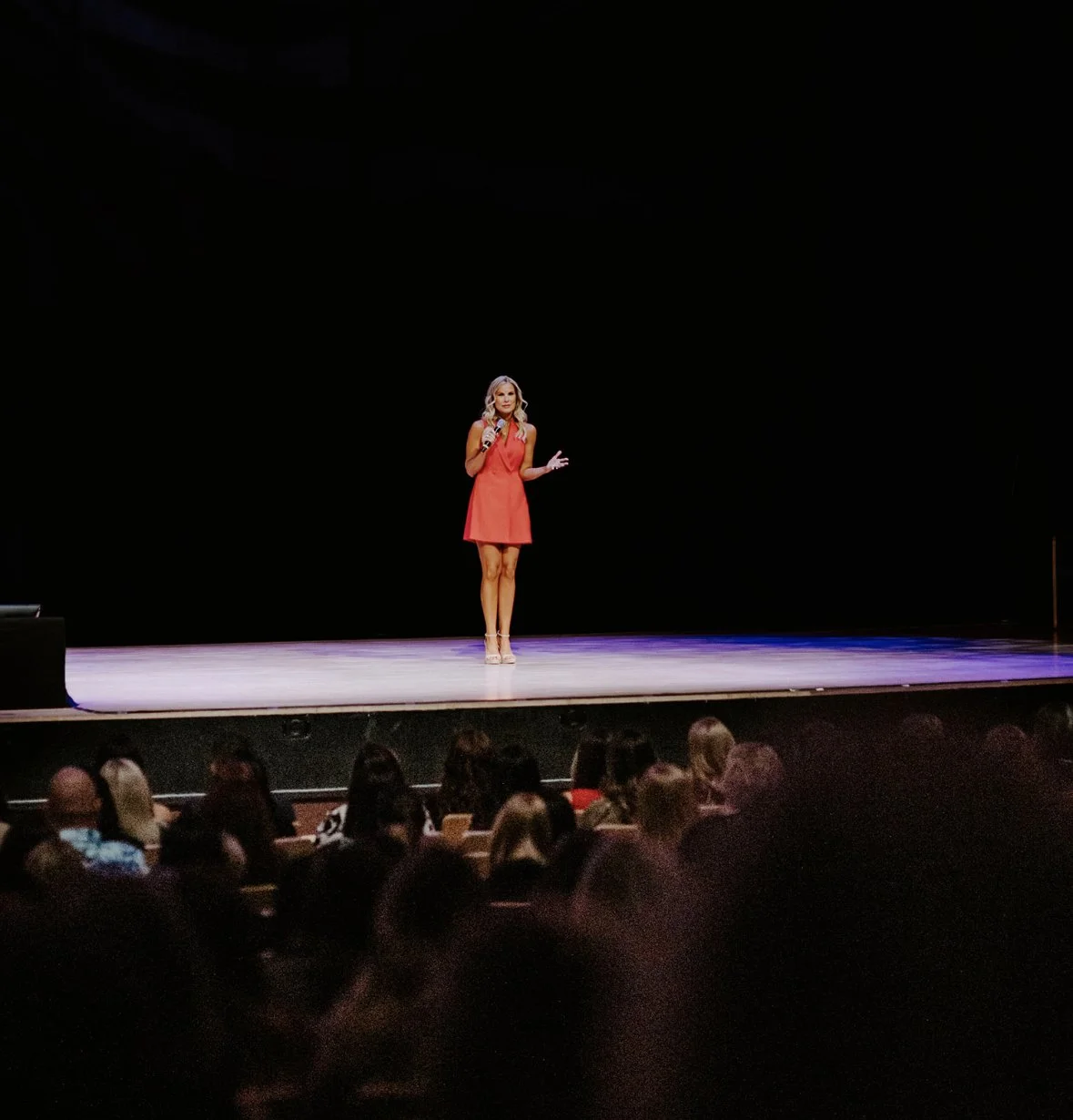 Woman in a red dress speaking into a microphone on a theater stage before an audience