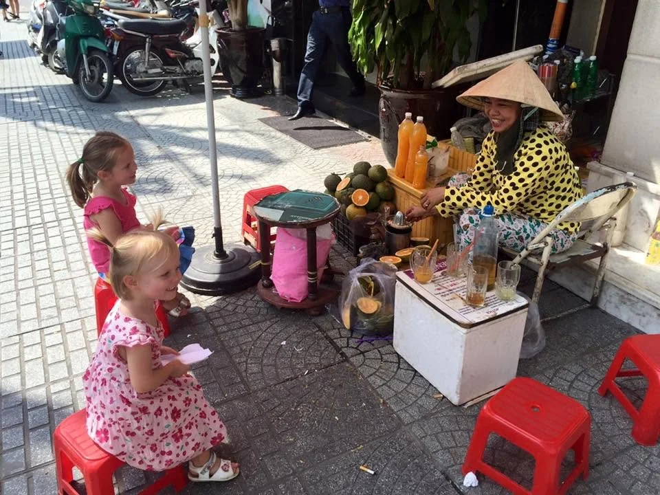 Vietnamese street vendor in a conical hat serving fresh orange juice to two young girls at a sidewalk fruit stand