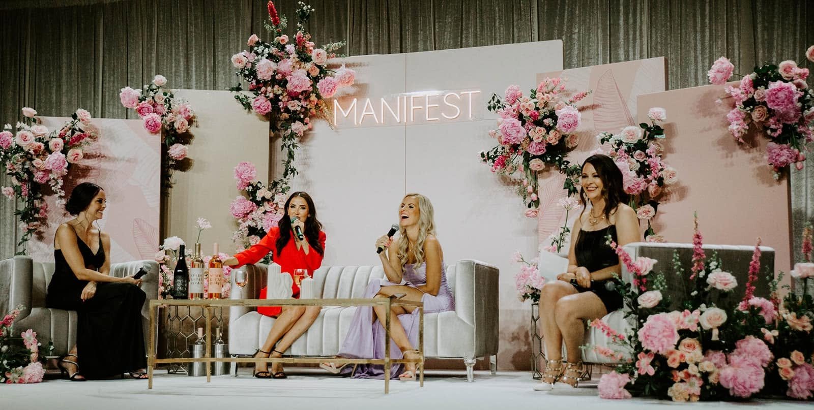 Four women speaking on a floral-decorated event stage beneath a glowing “MANIFEST” sign.