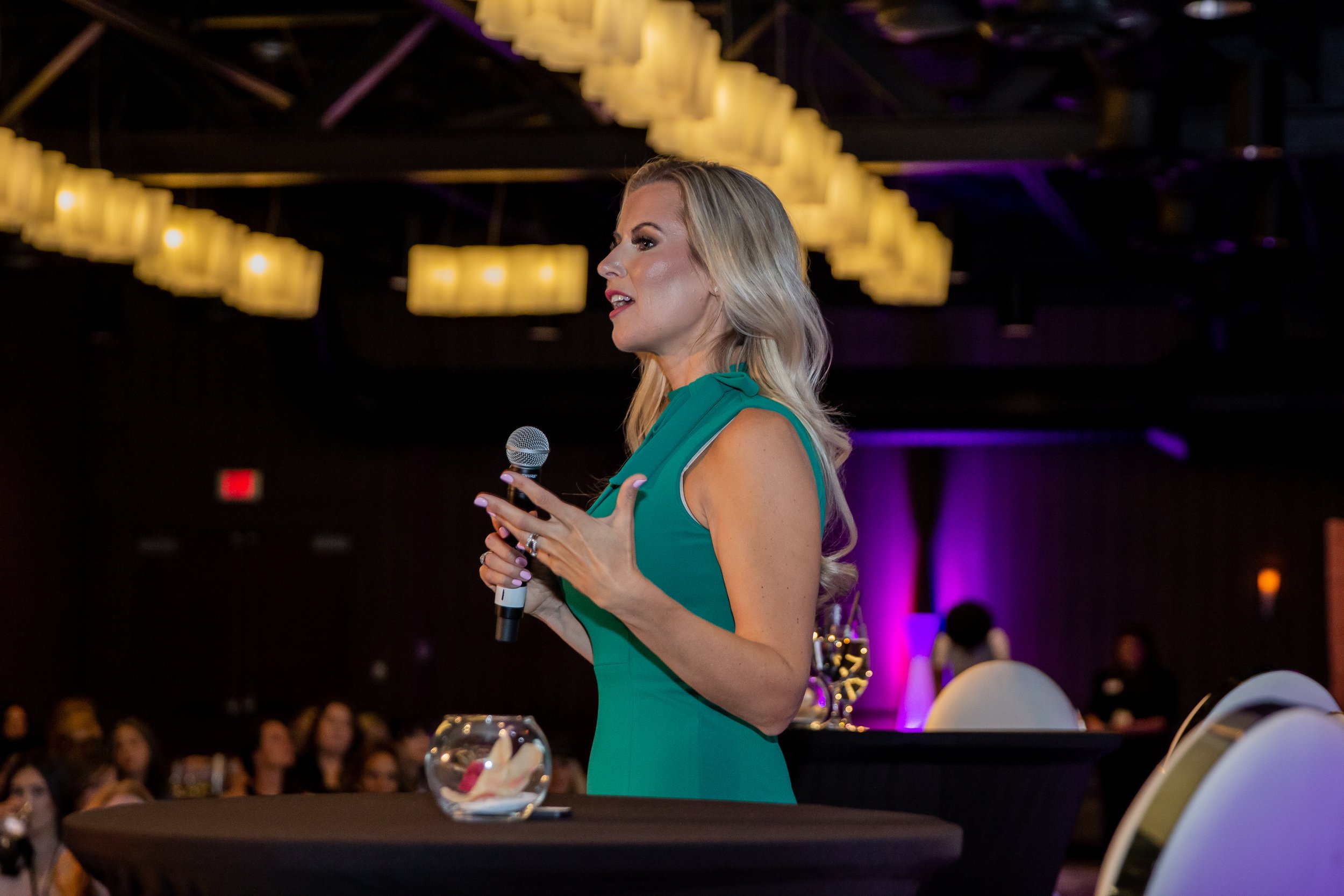 Blonde woman in a green dress speaking into a microphone at a dimly lit event with purple lighting.