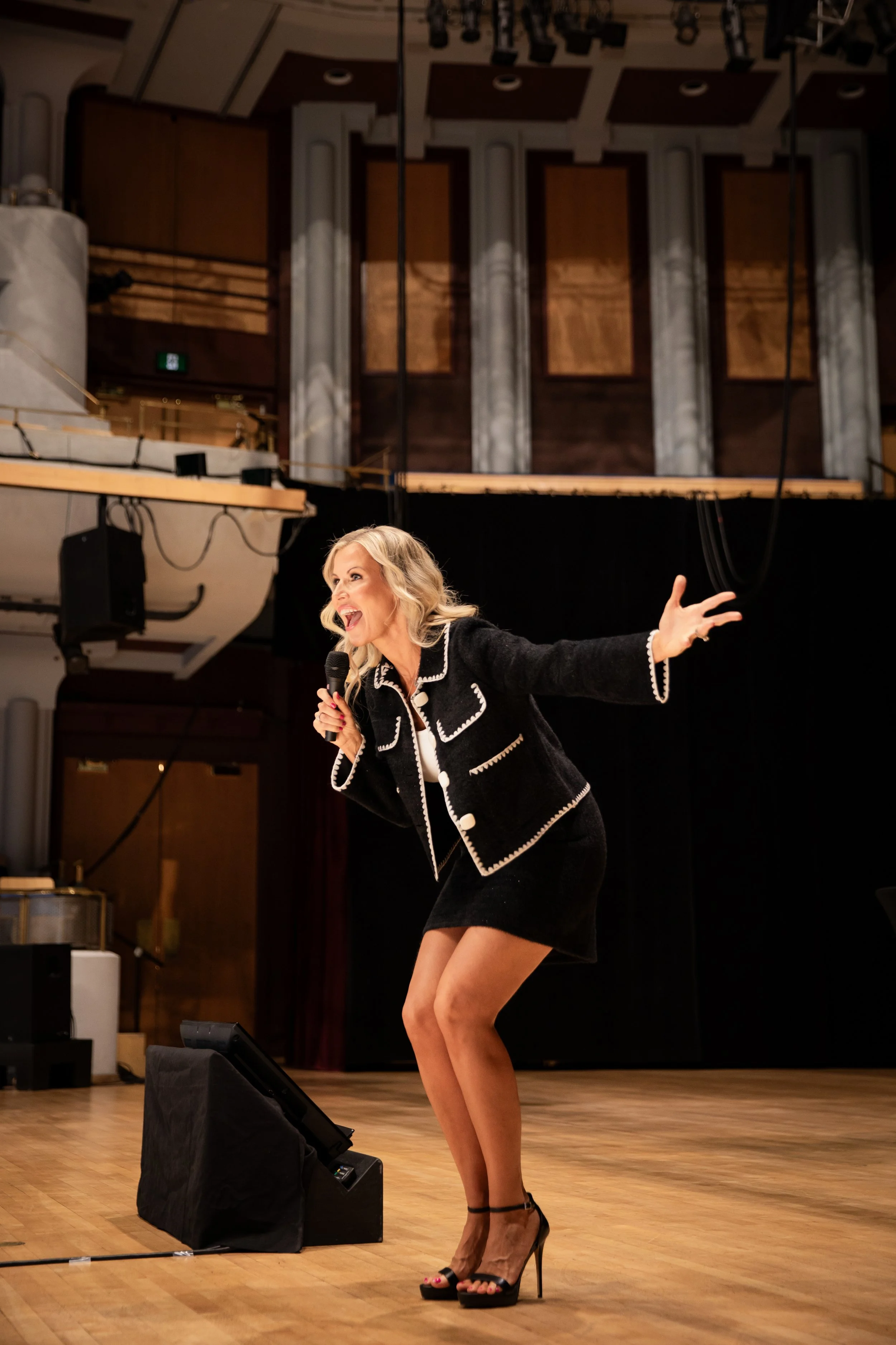 Blonde woman in a black-and-white outfit speaking into a microphone on a theater stage.