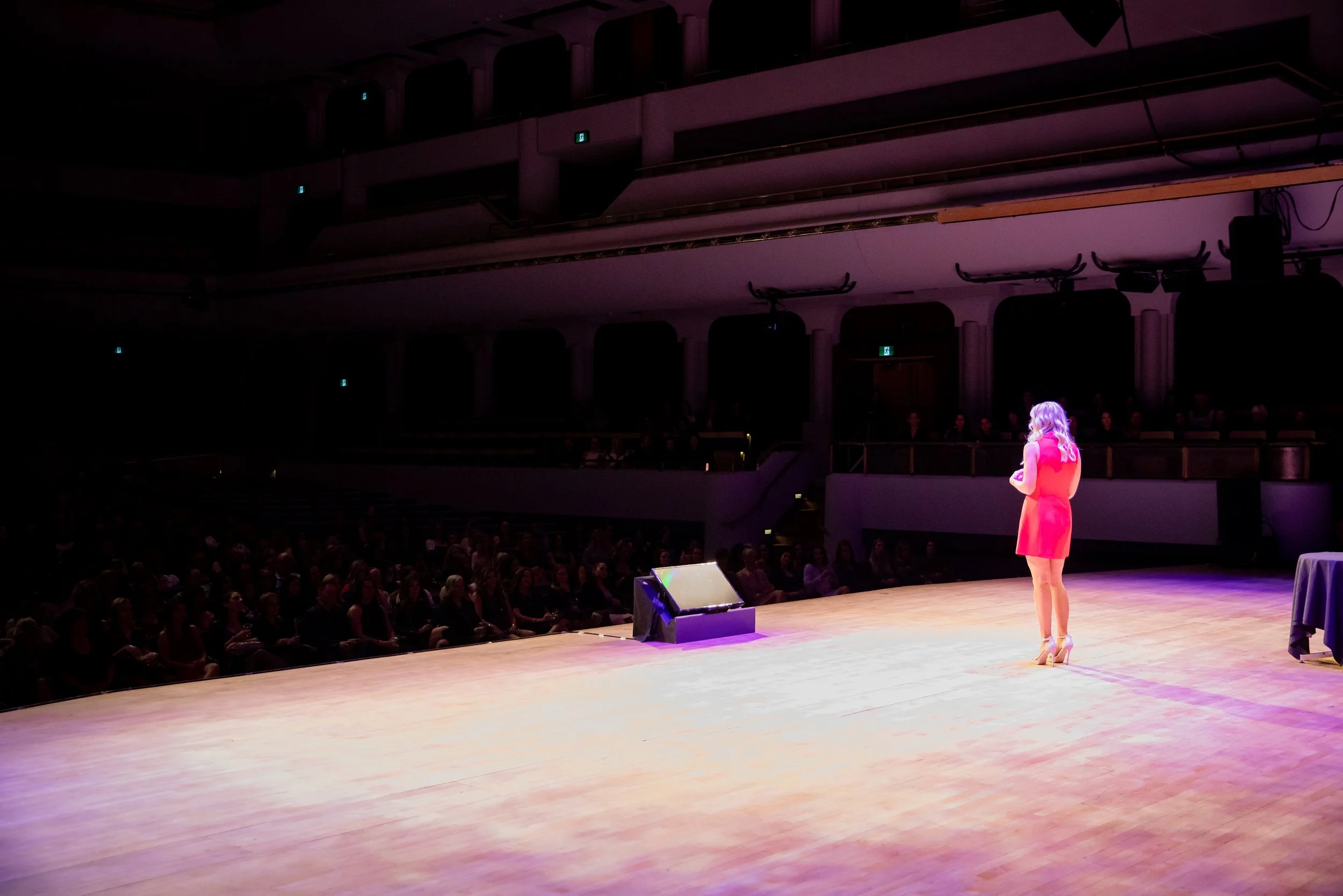 Woman in a bright pink dress speaking onstage to a seated audience in a theater
