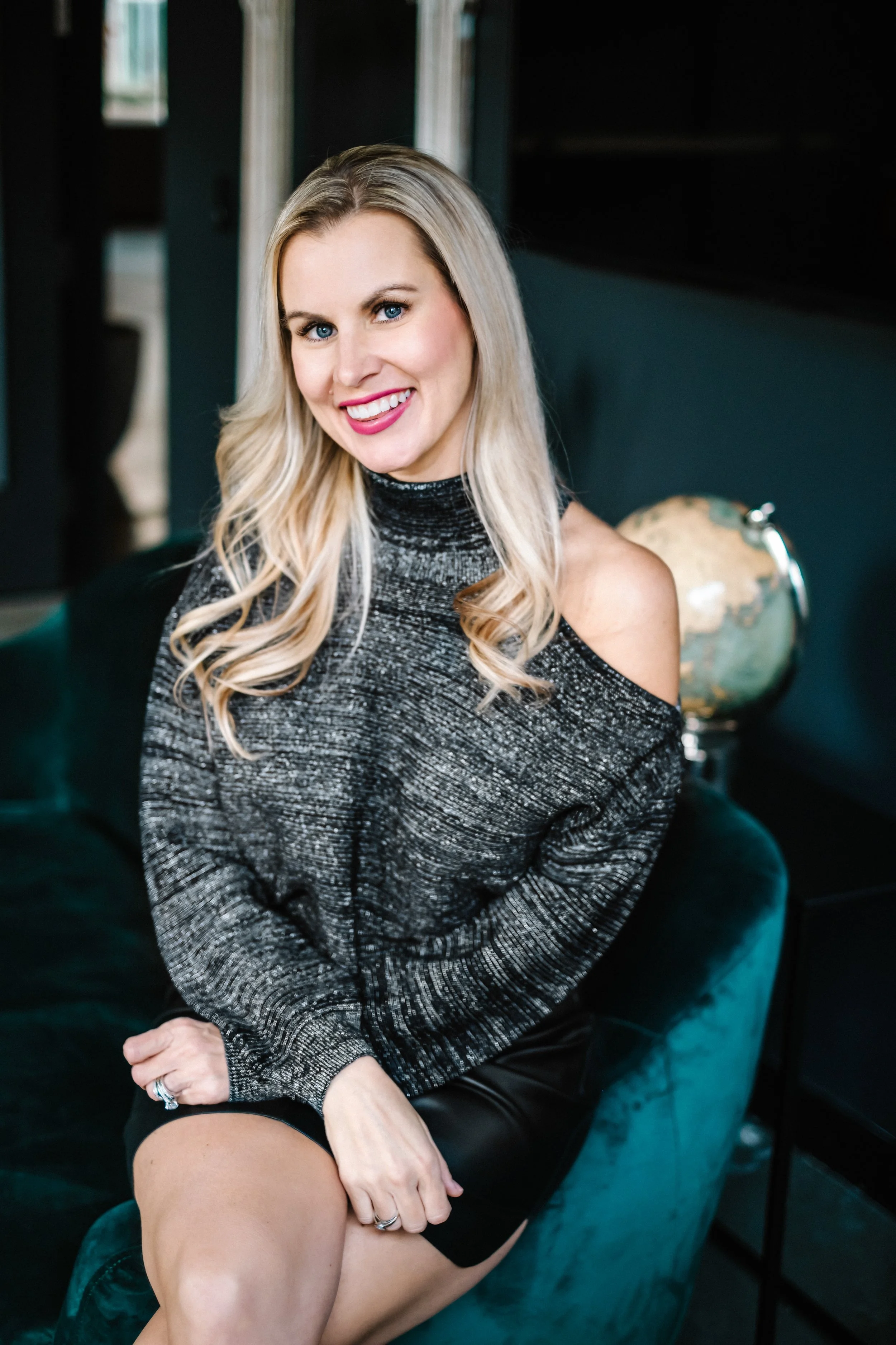 Smiling blonde woman with blue eyes sitting in a dark teal chair, wearing a sparkly gray sweater and black skirt.