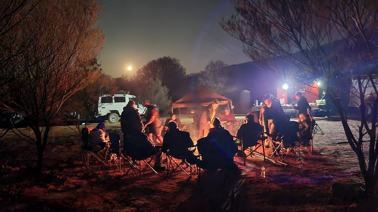 A group of people sitting around a campfire in Central Australia. There is a full moon in the background and the people with their backs to the camera are silhouetted in front of the campfire