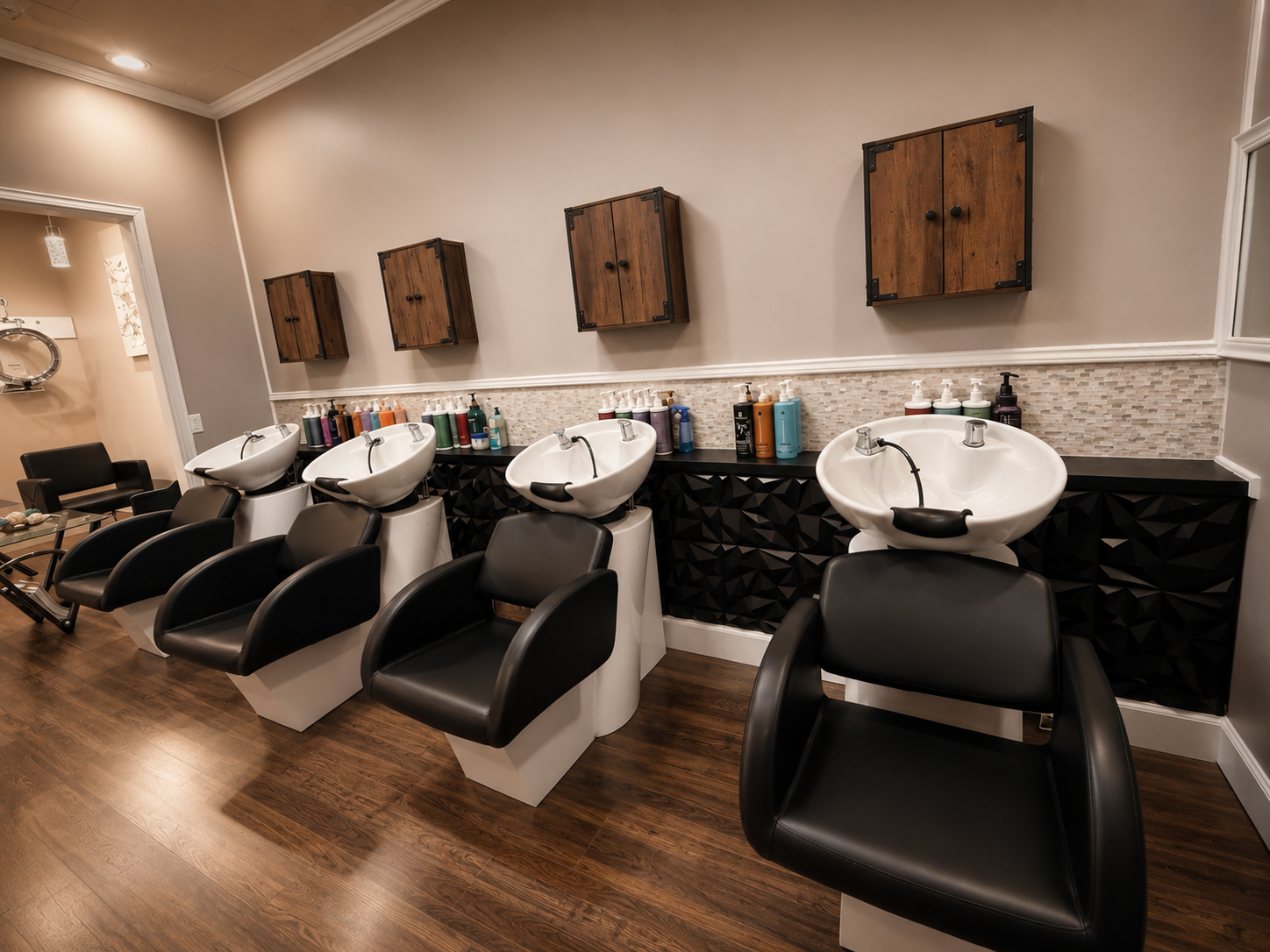 Salon with three white hair washing sinks, black chairs in front of them, wooden cabinets on beige wall, bottles of shampoo and conditioner, black and white decor.