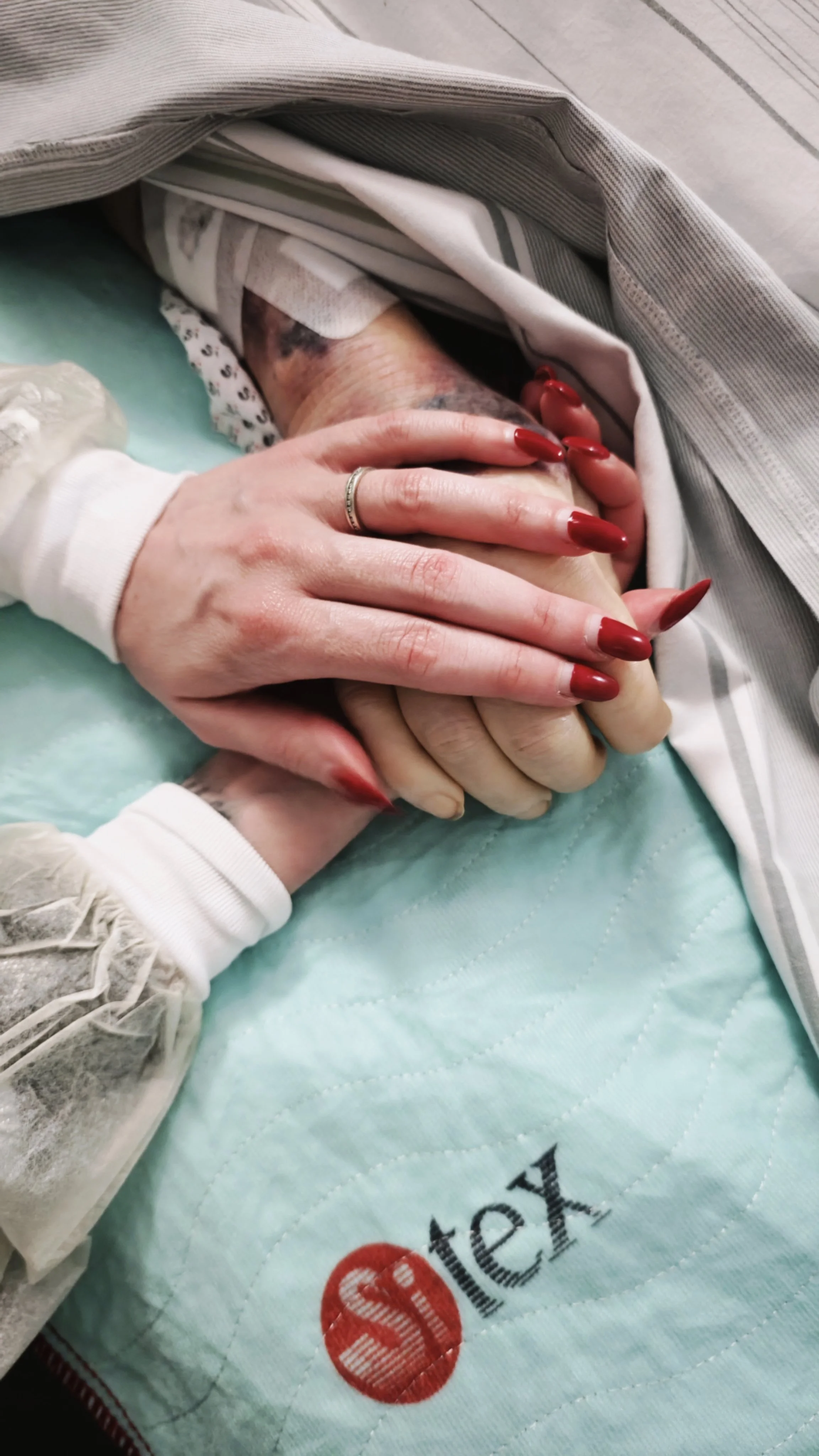 Two people with their hands clasped, one with red manicure and one with a wedding ring, resting on a hospital bed. An IV and medical bandages are visible, indicating a healthcare setting.