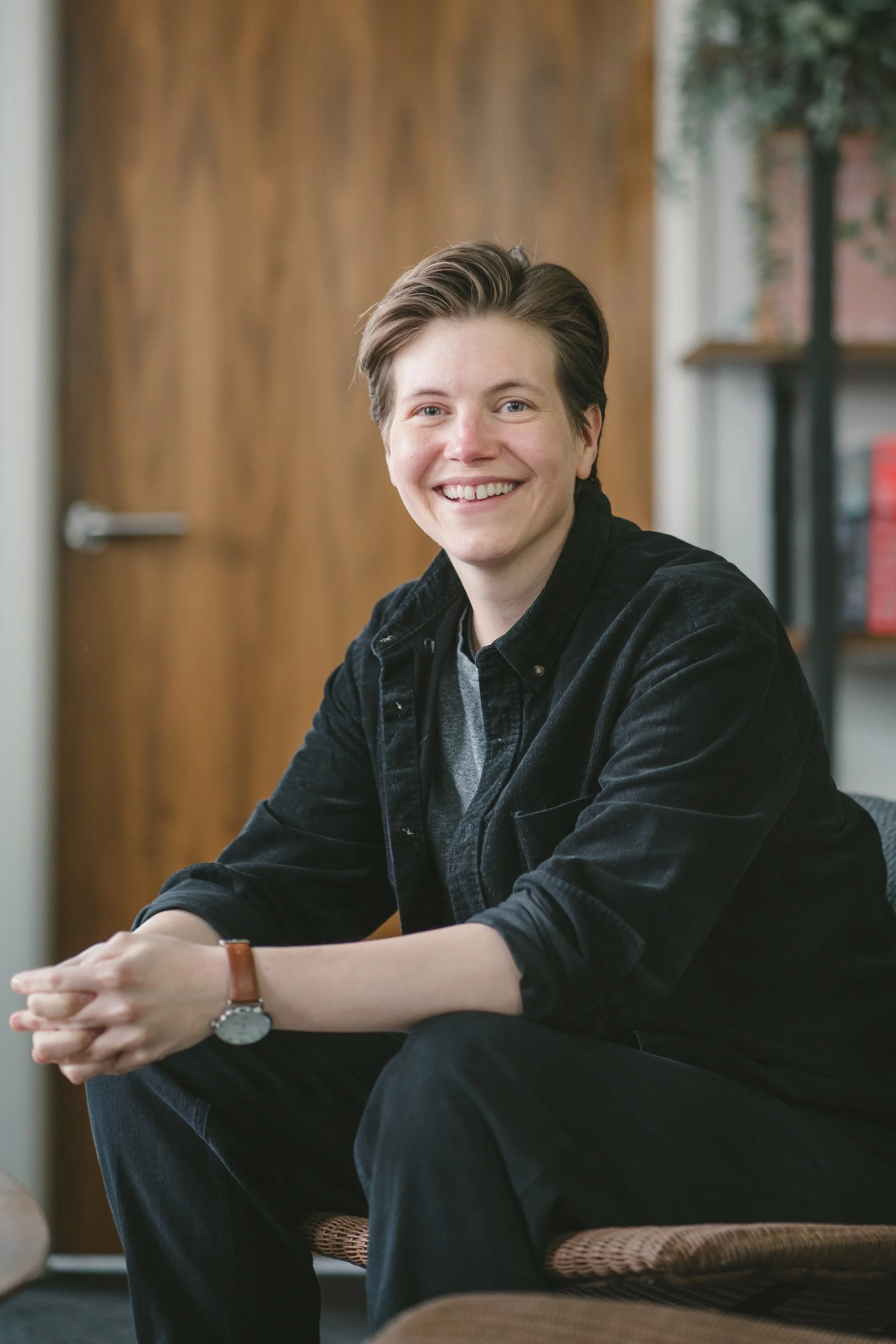 A young person with short hair smiling and sitting casually indoors with a wooden door and bookshelf in the background.