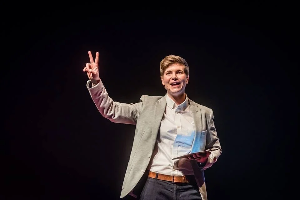 A person with short light brown hair, wearing a gray blazer and white shirt, stands against a black background, smiling and making a peace sign with their right hand while holding a tablet in their left hand.