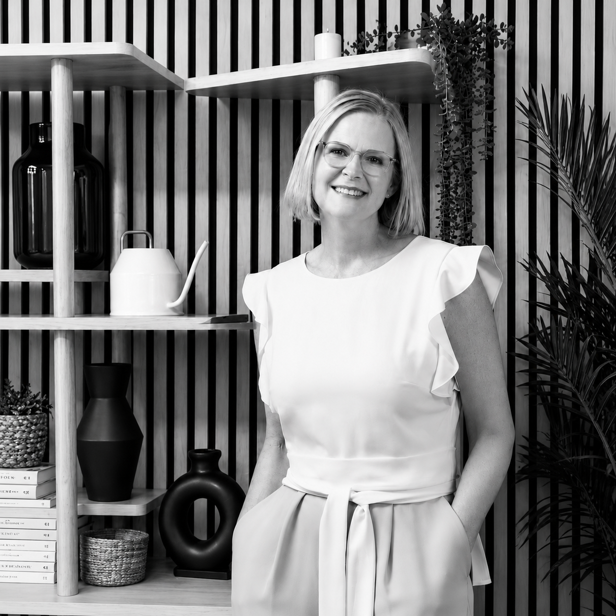 Black and white photo of a woman with short blonde hair and glasses smiling, standing in front of a wooden shelf with decorative vases, books, and plants.