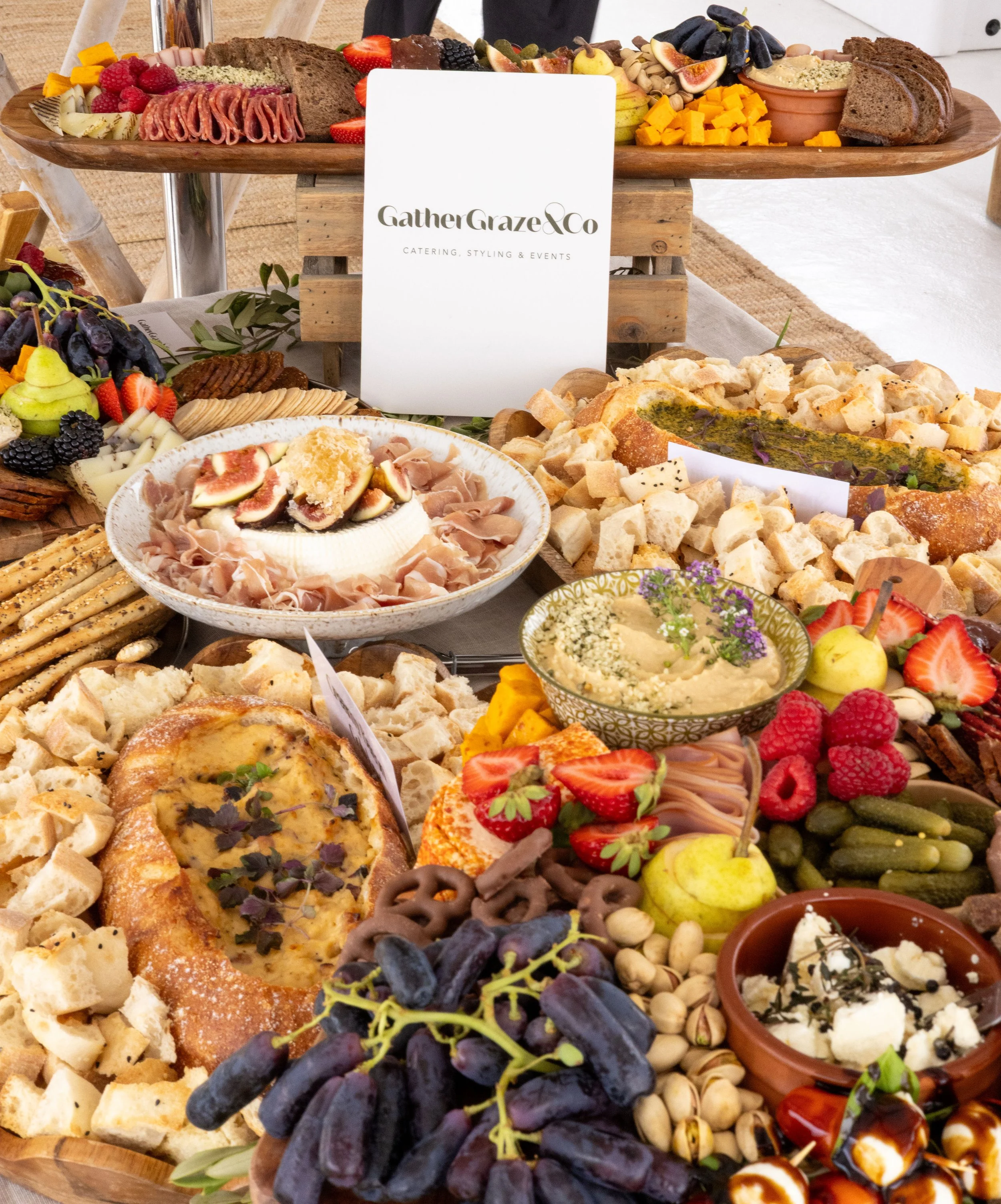 A large party platter of assorted cheeses, fruits, nuts, meats, and snacks with a stack of mini sandwiches, a layered chocolate cake with a "21" topper, and a floral arrangement on a wooden table.
