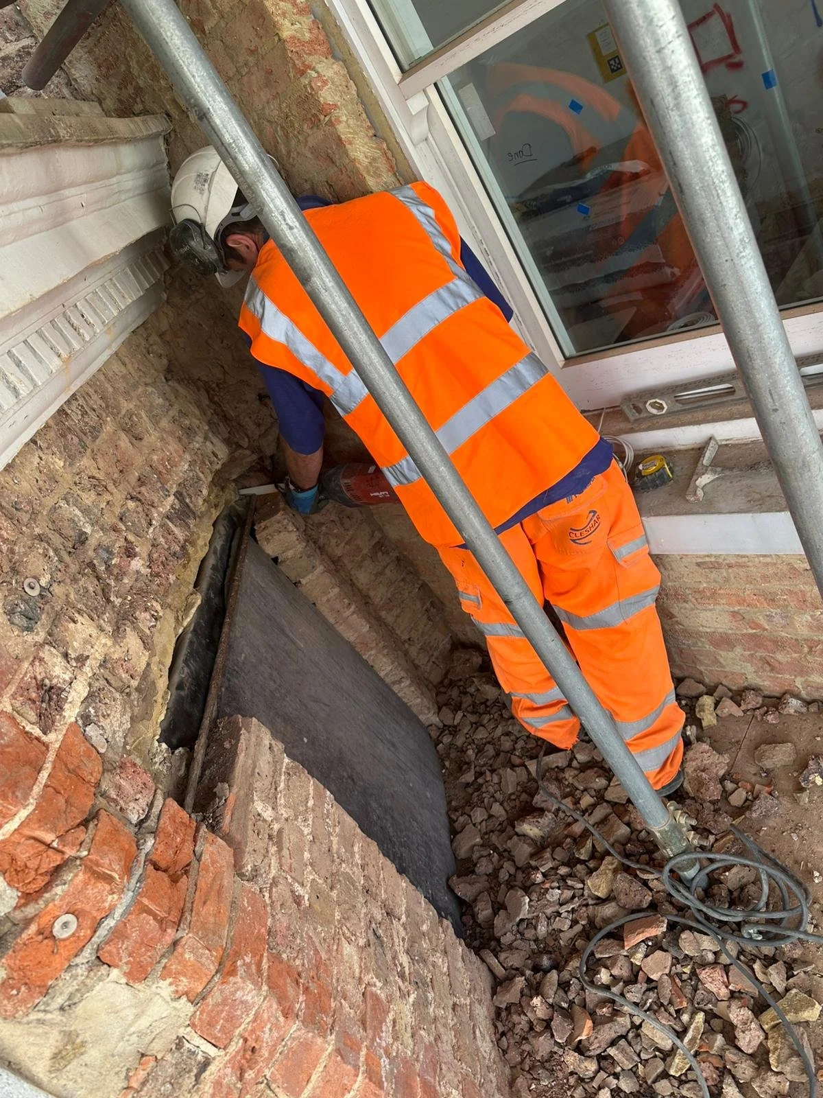 A construction worker wearing an orange safety vest and helmet is repairing a brick wall outside a building. The worker is focused on the wall and surrounded by construction debris, with metal scaffolding around him.