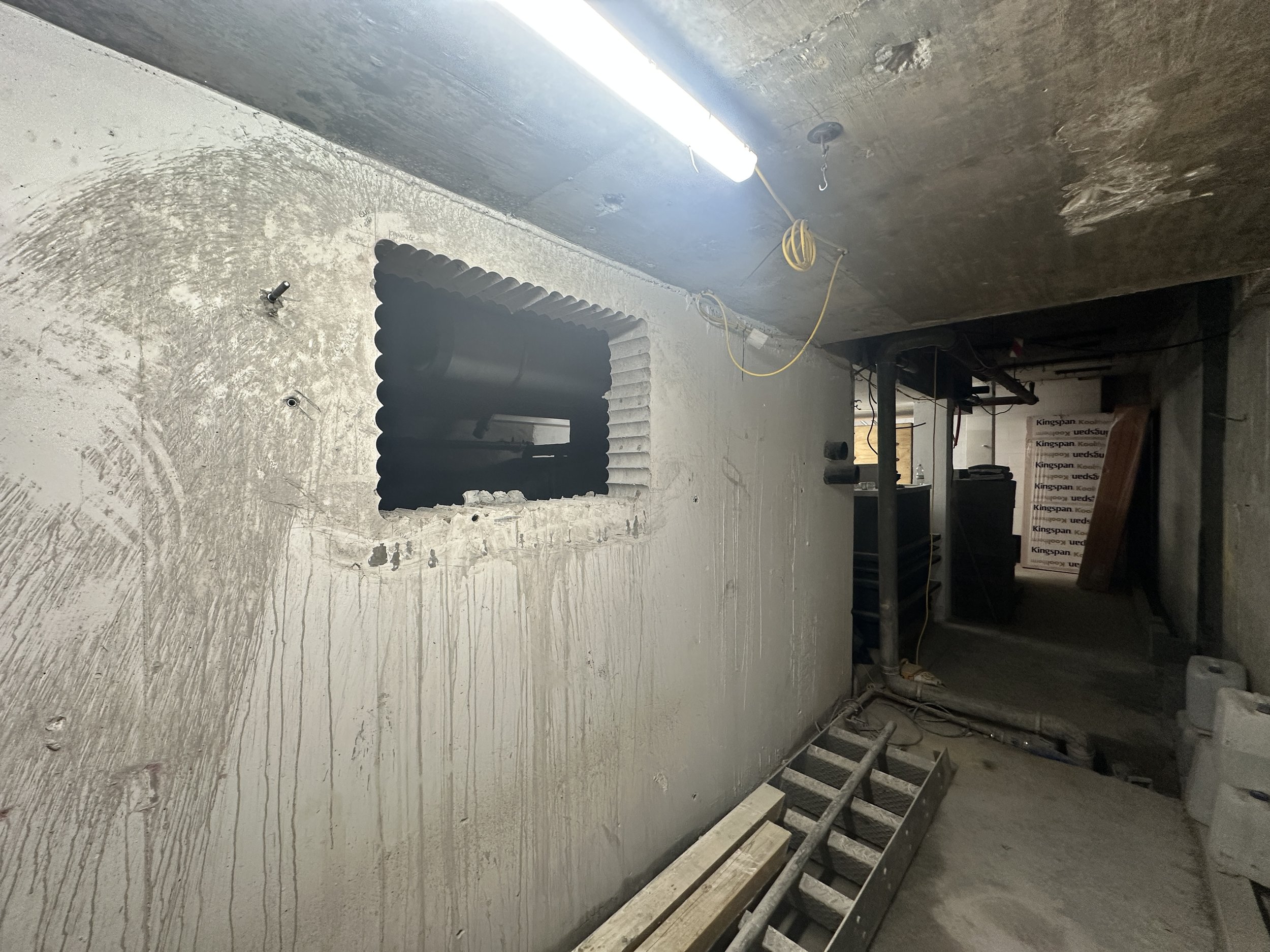 Construction site with unfinished concrete walls, a black corrugated pipe or duct, a fluorescent light, yellow electrical cords, and construction tools and materials.