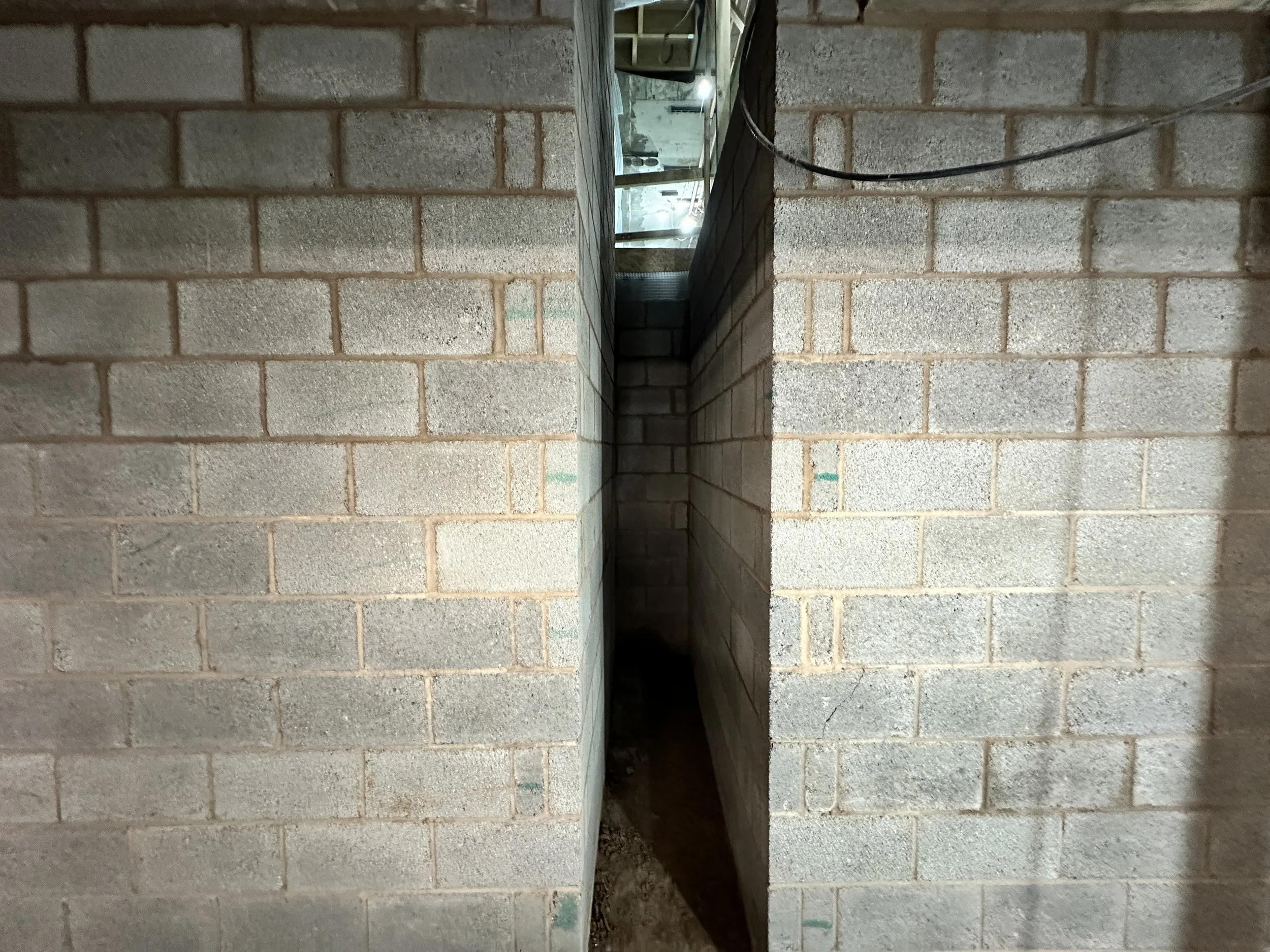 A narrow brick-walled alleyway with a covered sidewalk area. The image shows one side of the alley with beige bricks and some green markings, and the other side with darker bricks. The alley appears to be in an unfinished or under-construction building, with exposed wiring and structural elements visible at the far end.