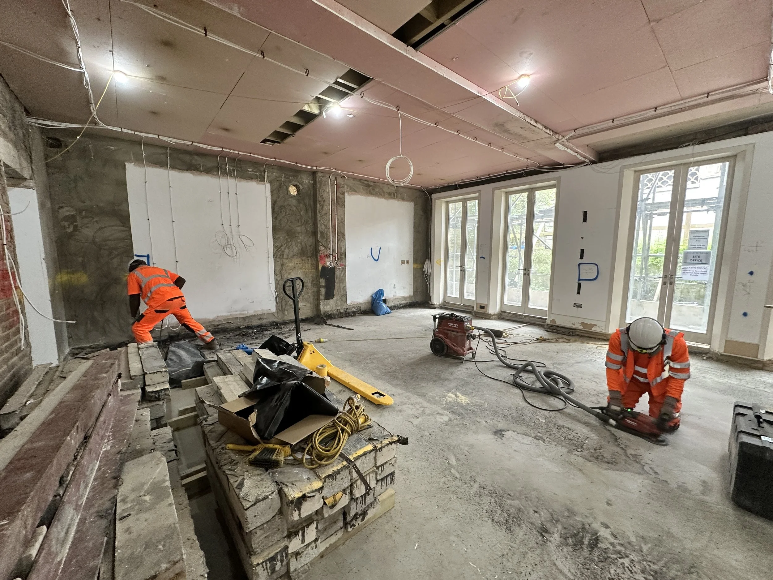 Construction workers in orange safety clothing working inside a building under renovation, with exposed wiring on the ceiling and large windows letting in natural light.