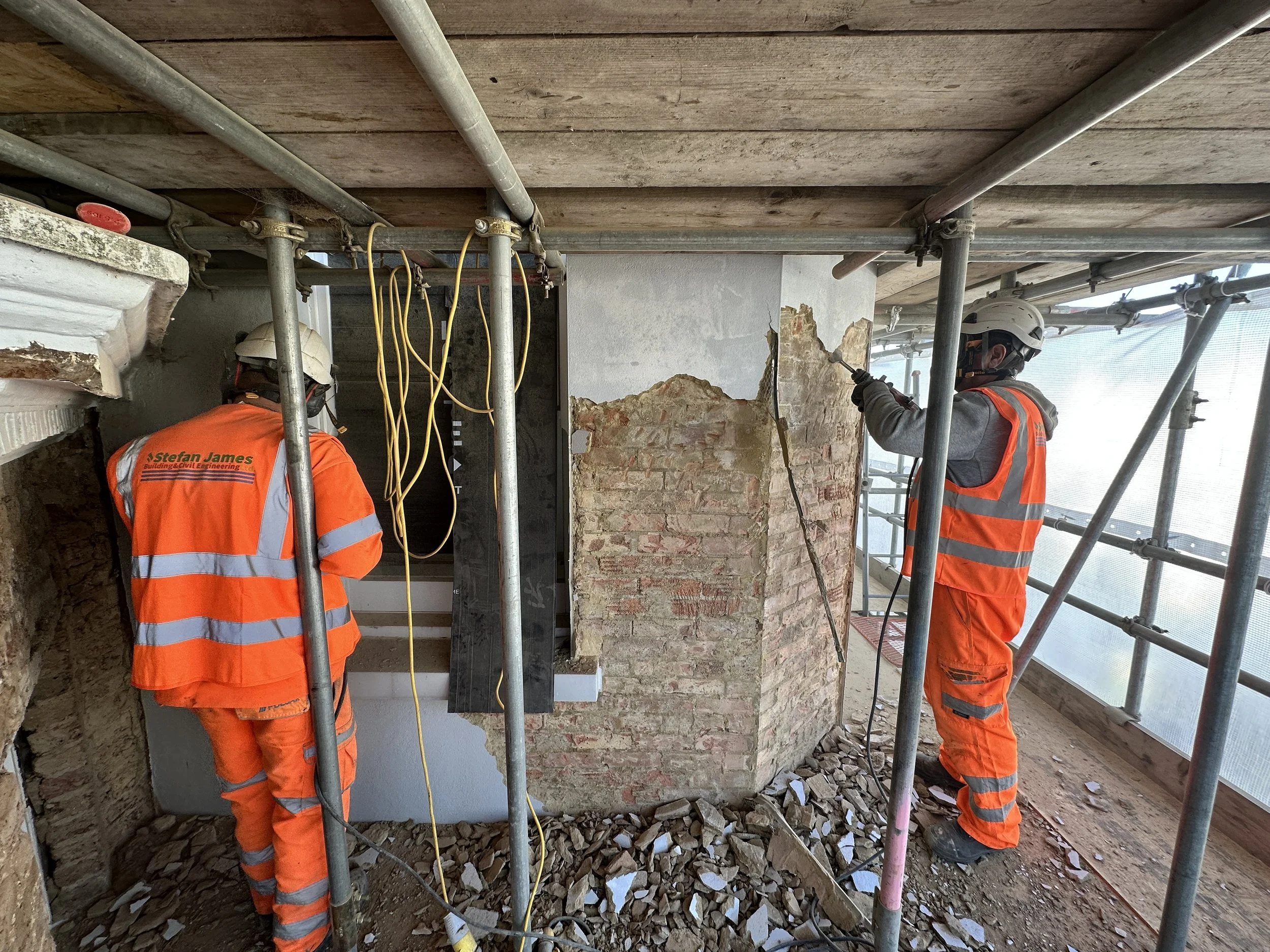 Two construction workers wearing orange safety vests and helmets working on a building renovation. One is using a power tool on brick wall, scaffolding surrounds them, and debris is on the ground.