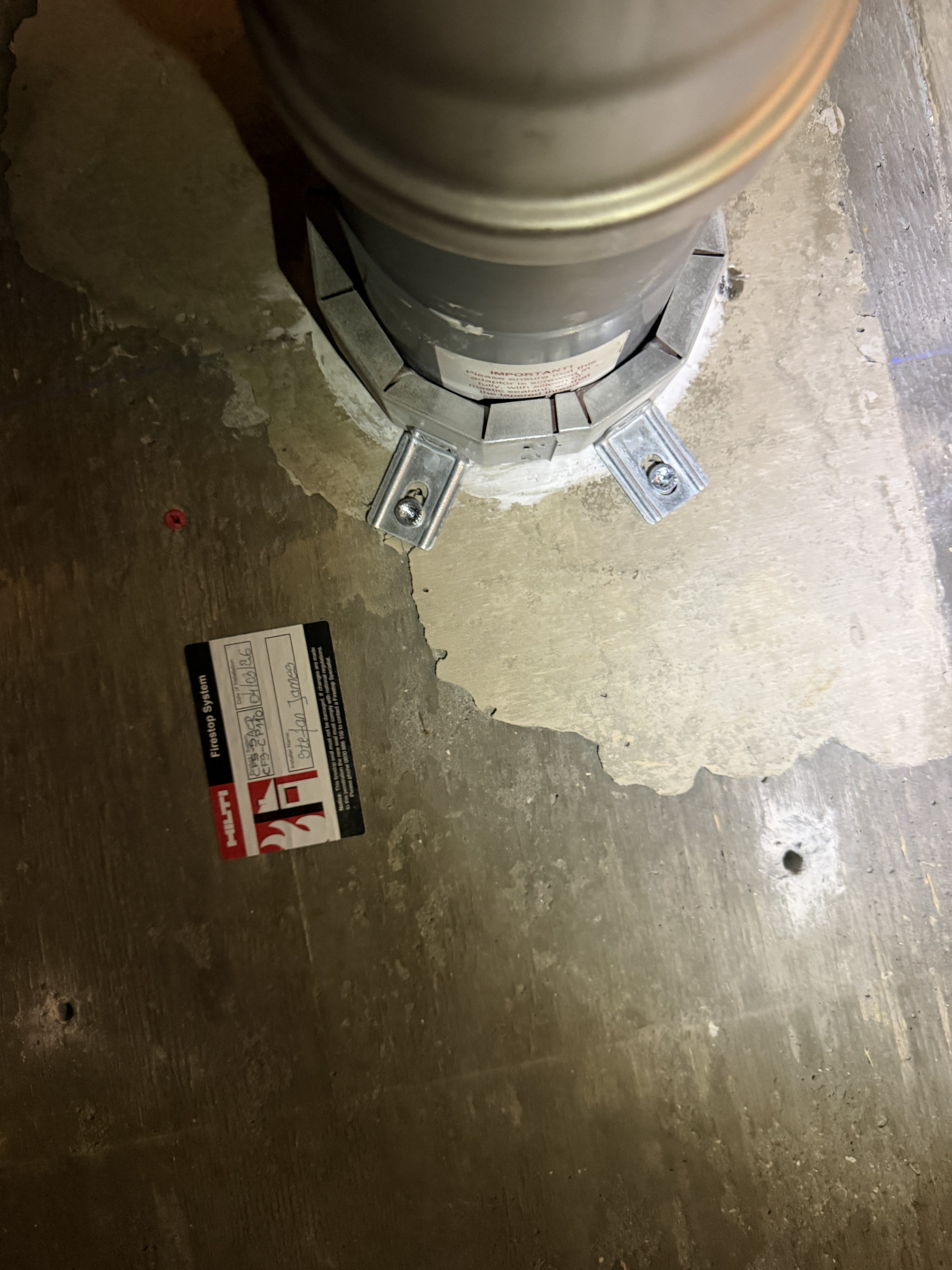 Close-up of a building's foundation with a metal post anchored to the concrete floor using metal brackets and bolts, with a warning sticker on the post and some construction materials nearby.