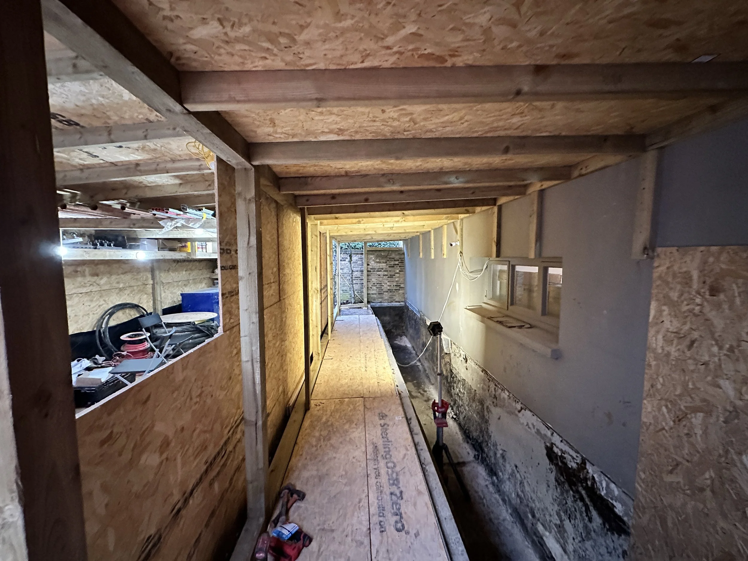 Construction site interior showing unfinished wooden framework, partially installed drywall, construction tools, and materials.
