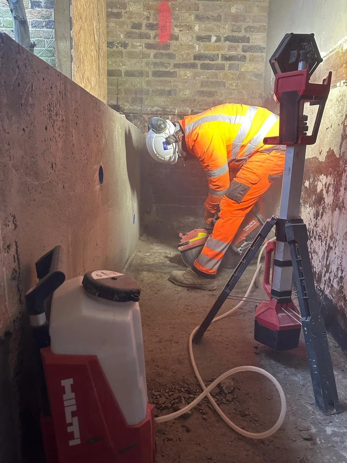 Construction worker wearing an orange safety suit and helmet using a power tool while working in a narrow, unfinished brick room.