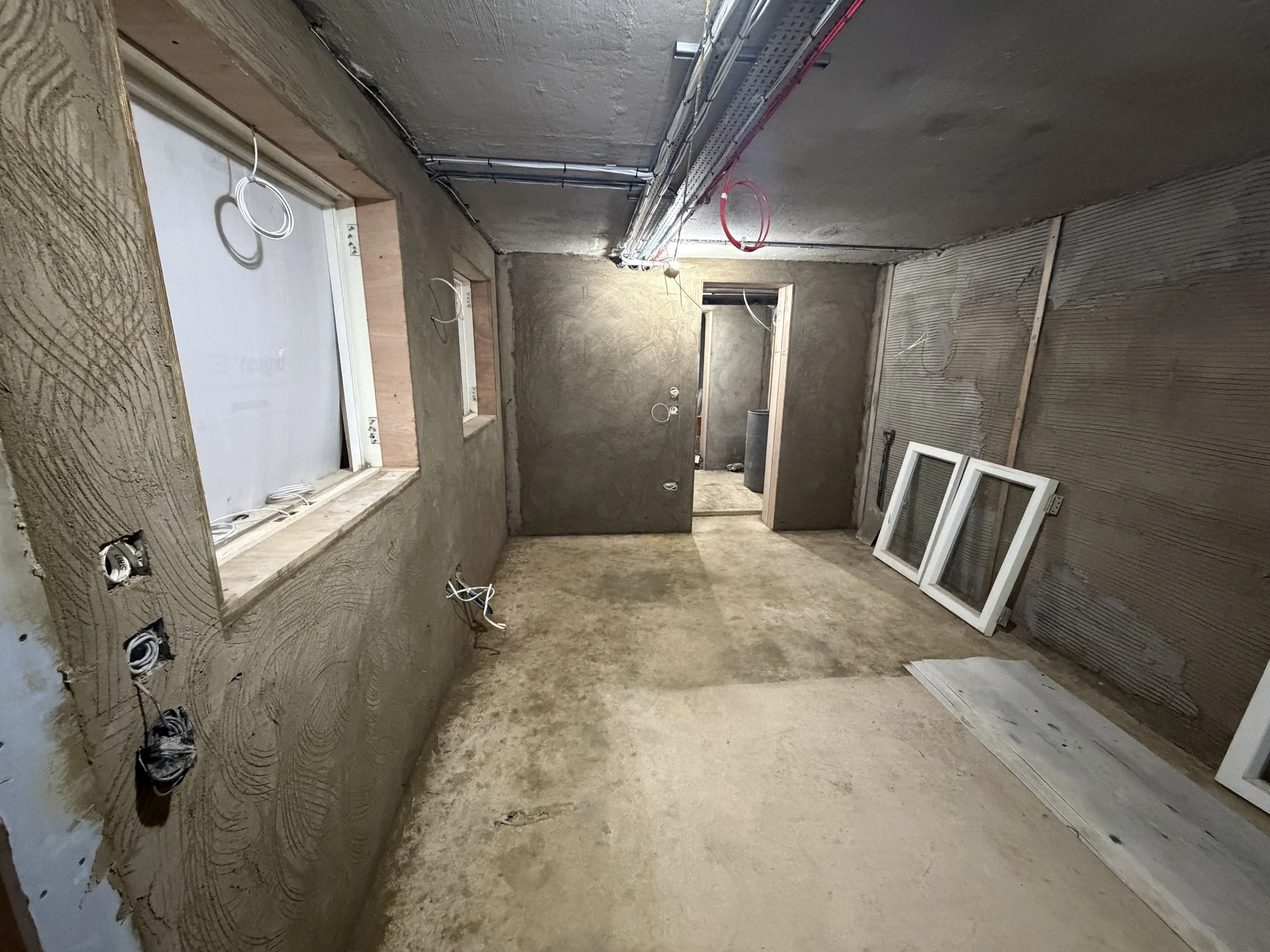 Interior of a building under construction or renovation, showing exposed wiring, unfinished walls, and window frames leaning against the wall.