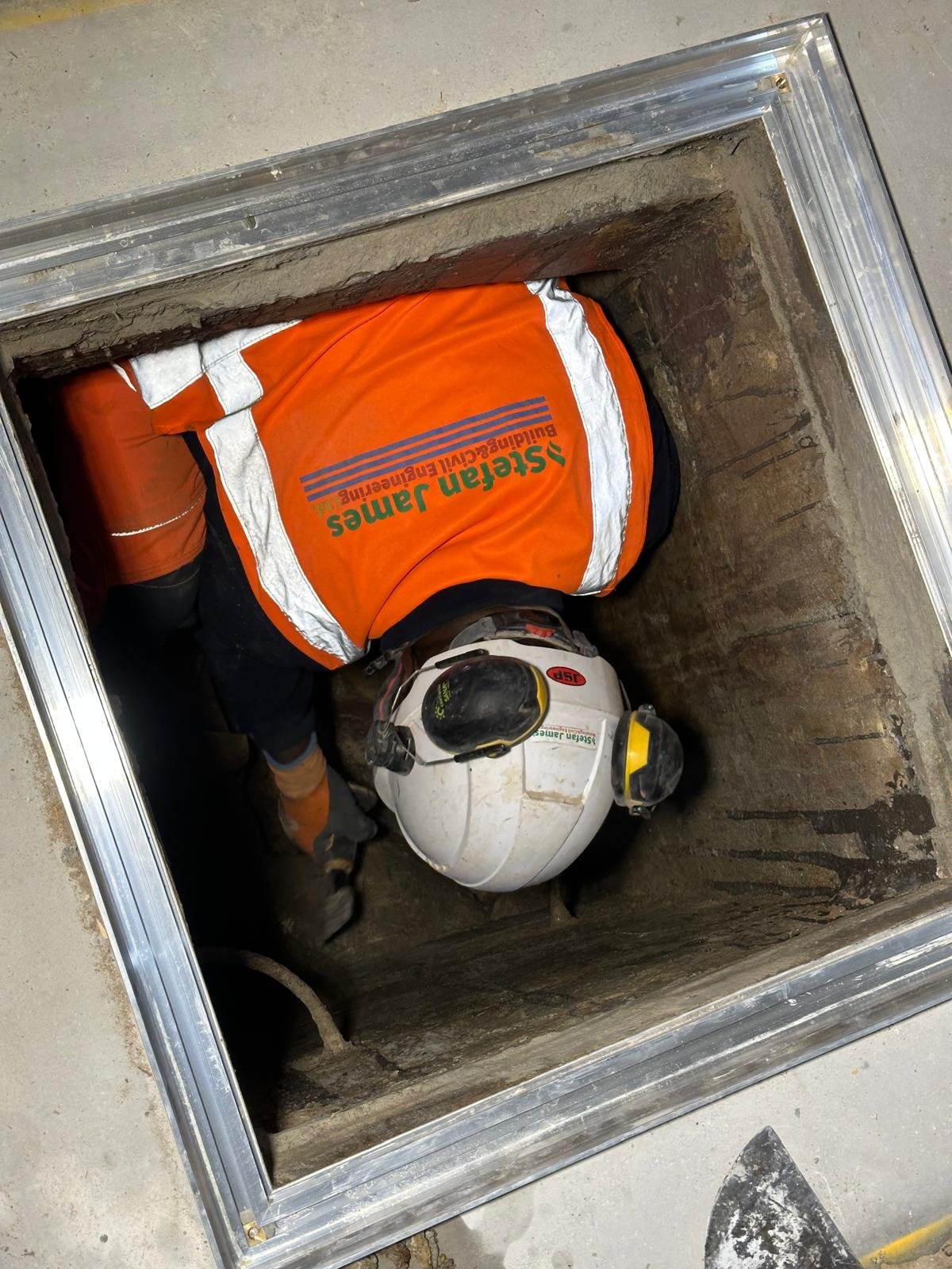 A worker crawling into an underground maintenance or inspection hole, wearing an orange vest with the logo of Steam Jam and a helmet, in a construction or infrastructure setting.