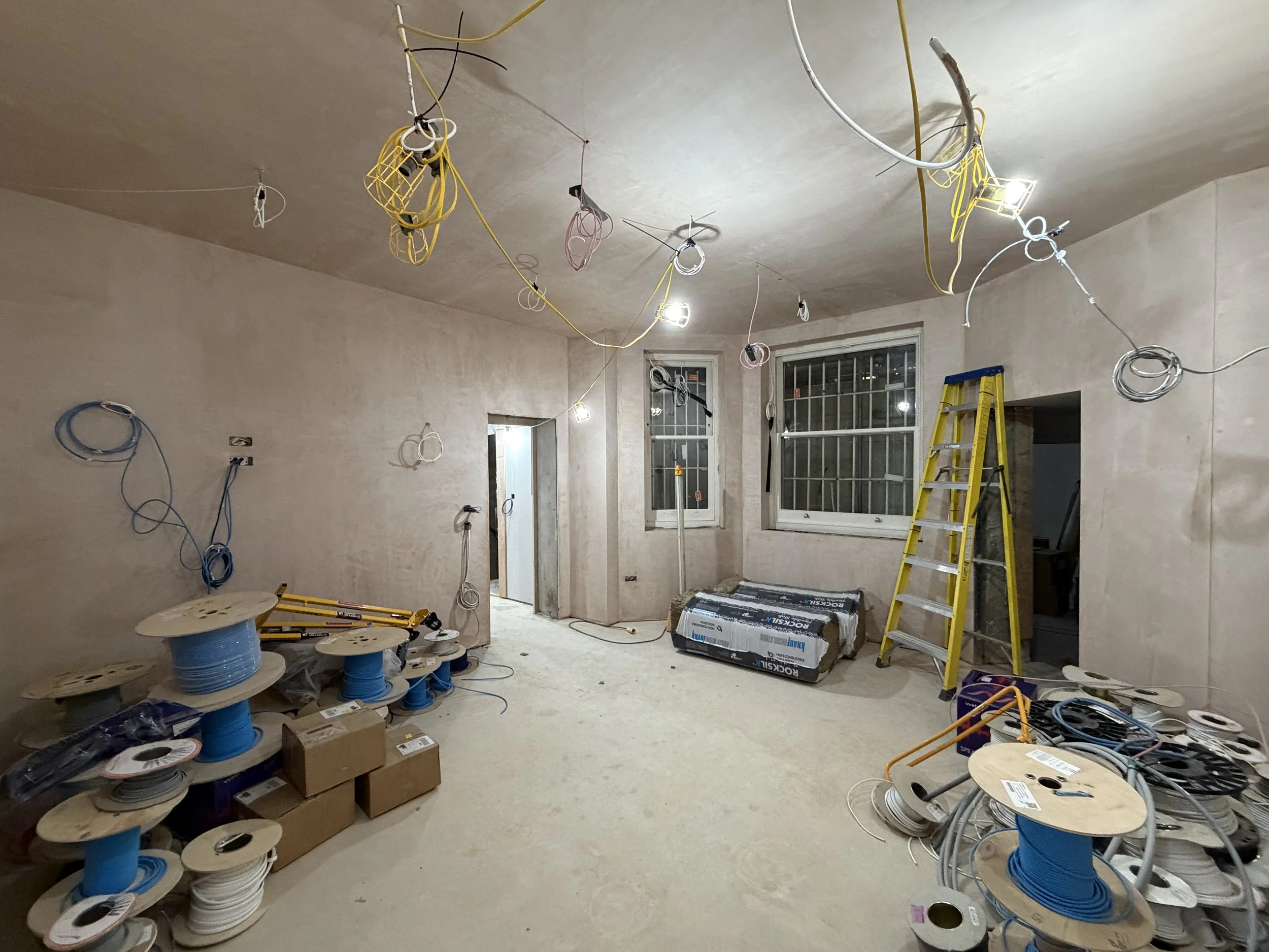 Room under construction or renovation with wires, tools, and equipment. Yellow ladder, window, and various spools of electrical wire are visible.