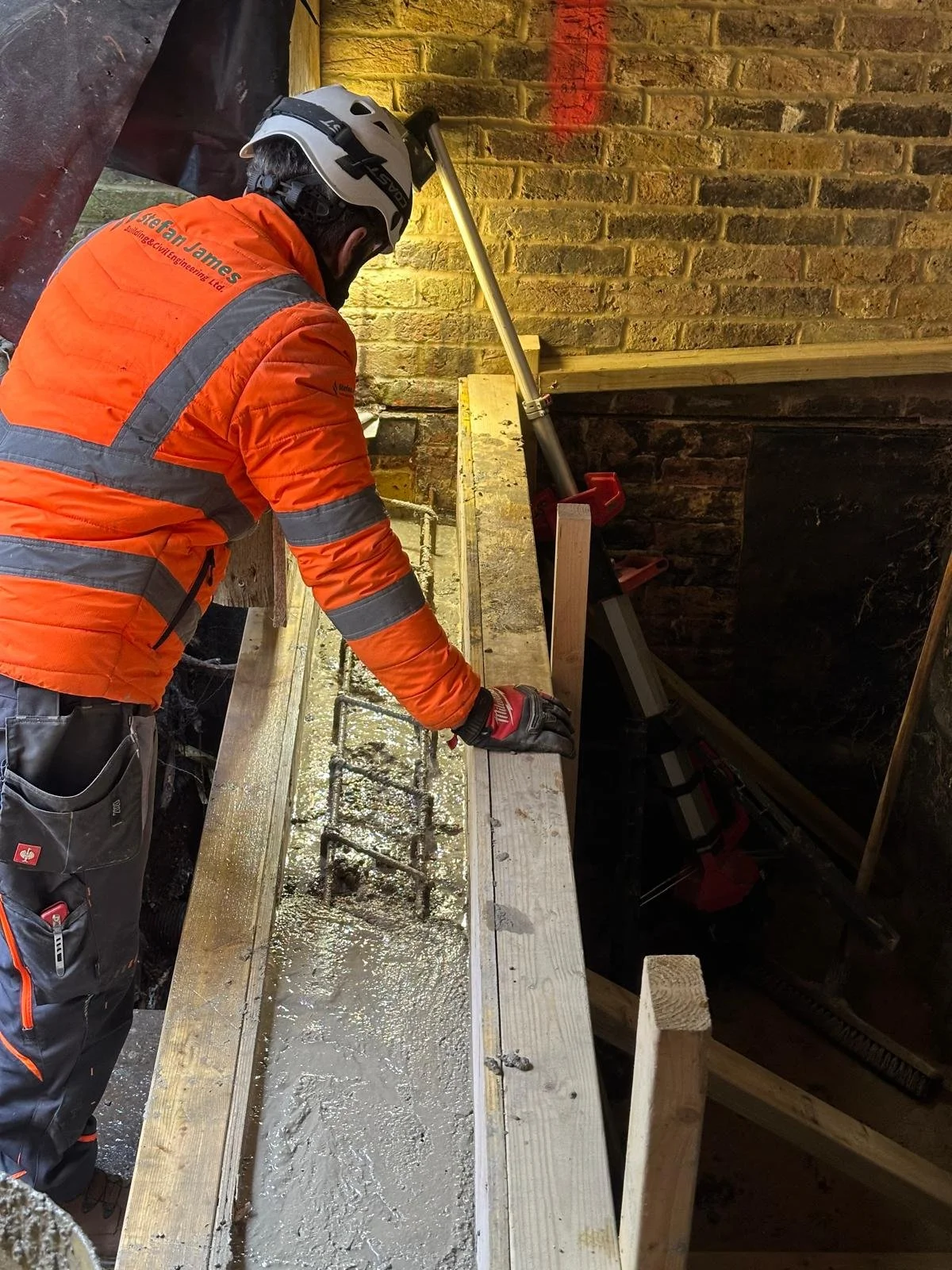 A construction worker wearing an orange safety jacket and a helmet is working on a brick wall, smoothing wet concrete on a wooden form. The worker is using a trowel and is dressed in gloves and safety gear.