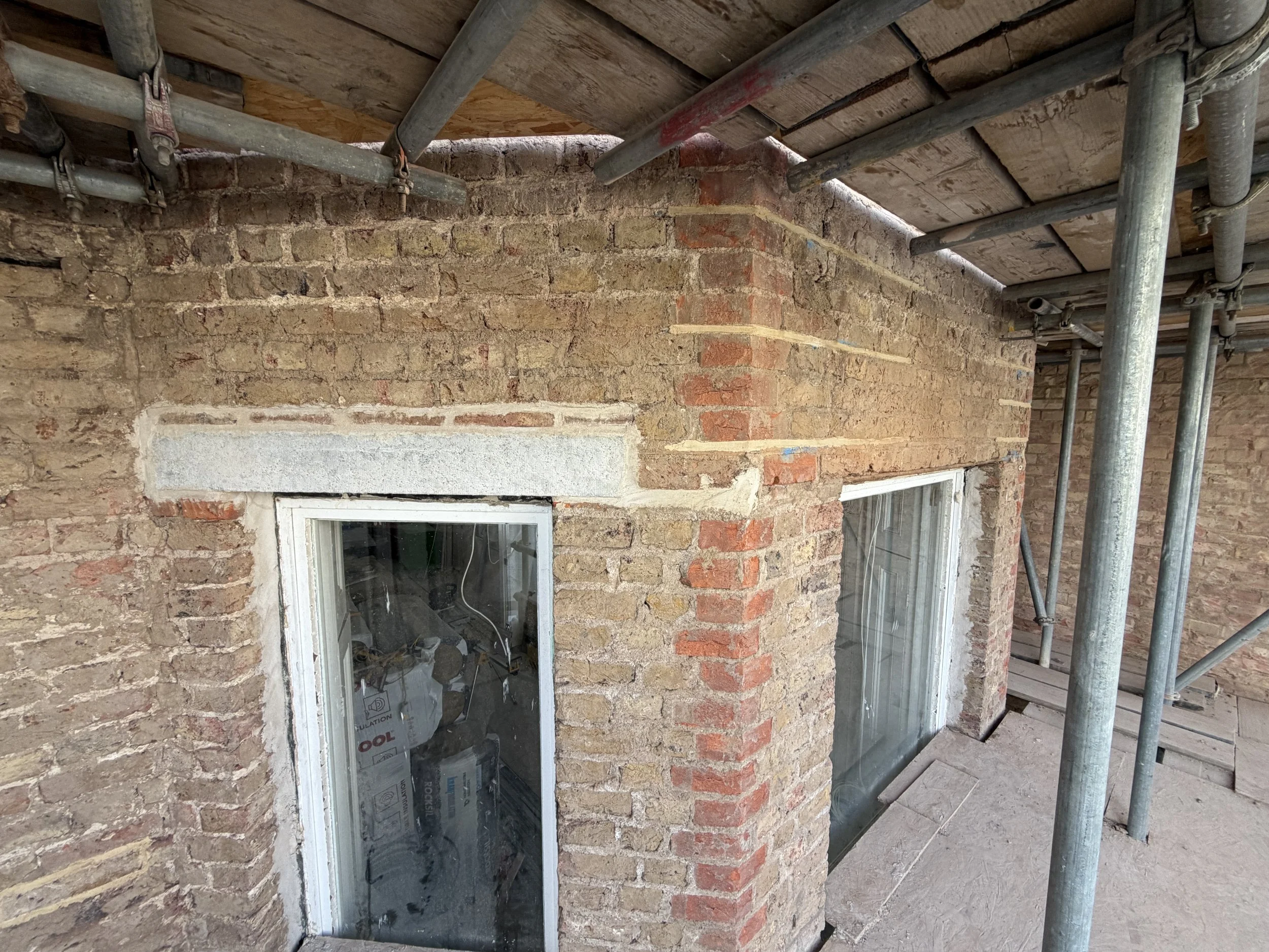 A building under construction with brick walls, two windows, and scaffolding on the exterior. The roof is supported by pipes and wooden planks.