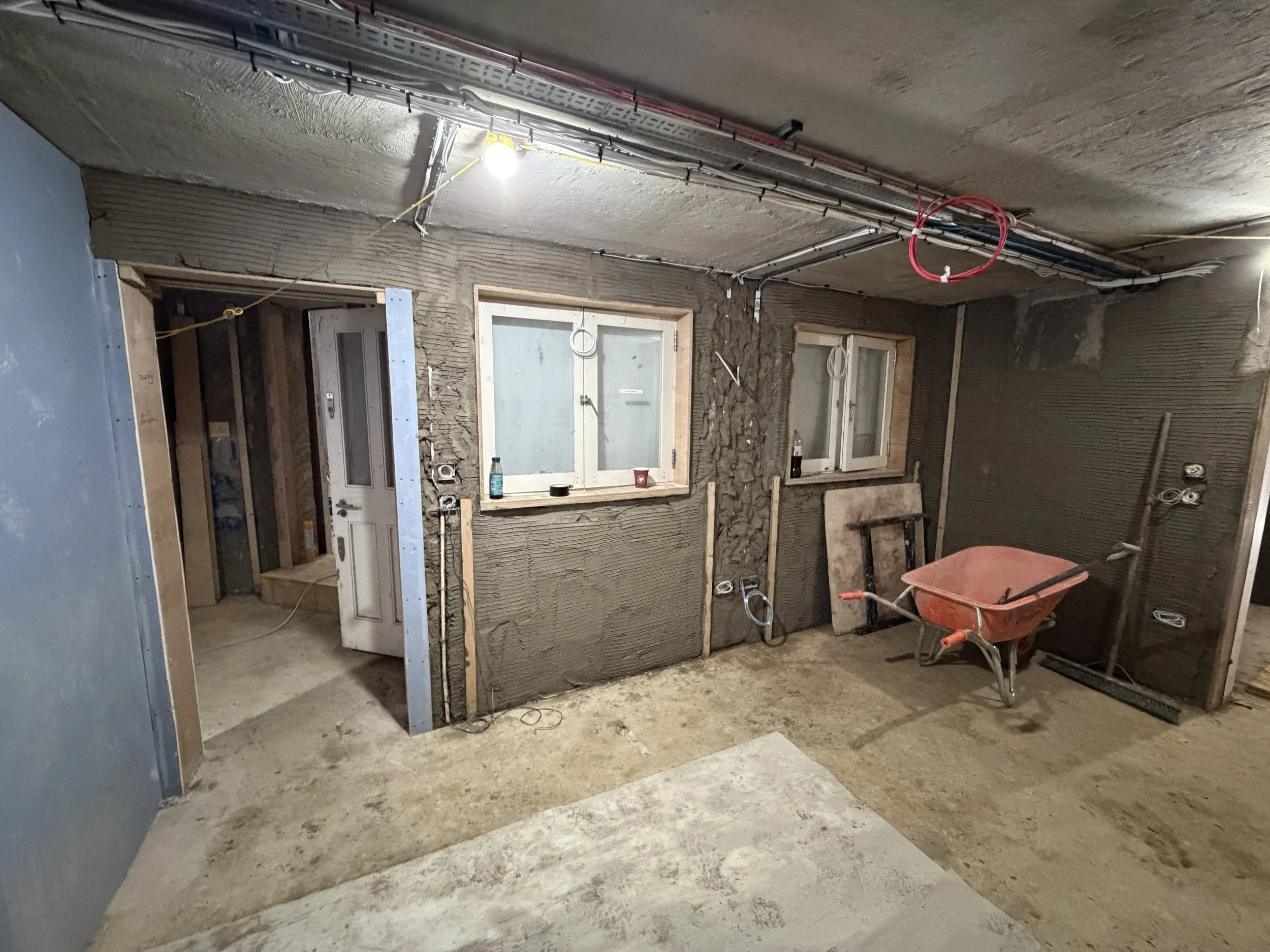 An unfinished interior room under construction with exposed concrete and wiring. There are two windows with wooden frames, some construction tools including a red wheelbarrow, and construction materials scattered around.