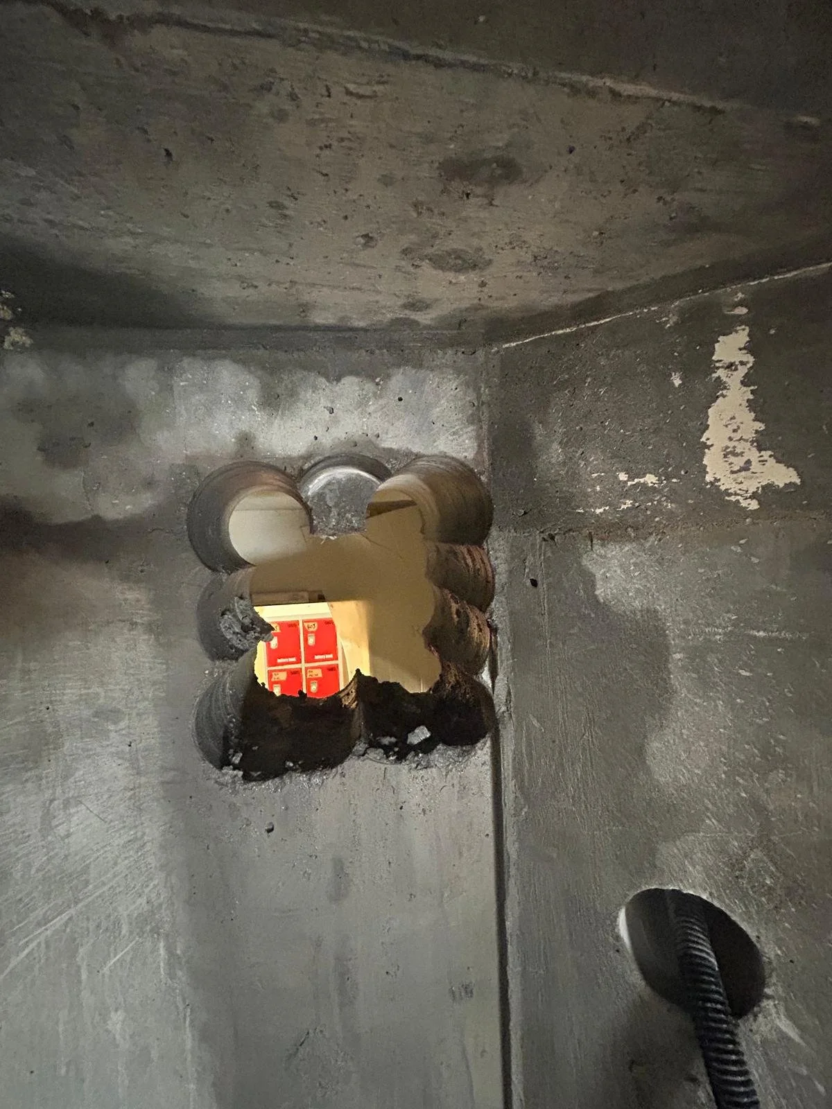 A hole in a concrete wall with several concrete pieces broken off around it, revealing a view of a fire alarm panel with red background and white labels inside.