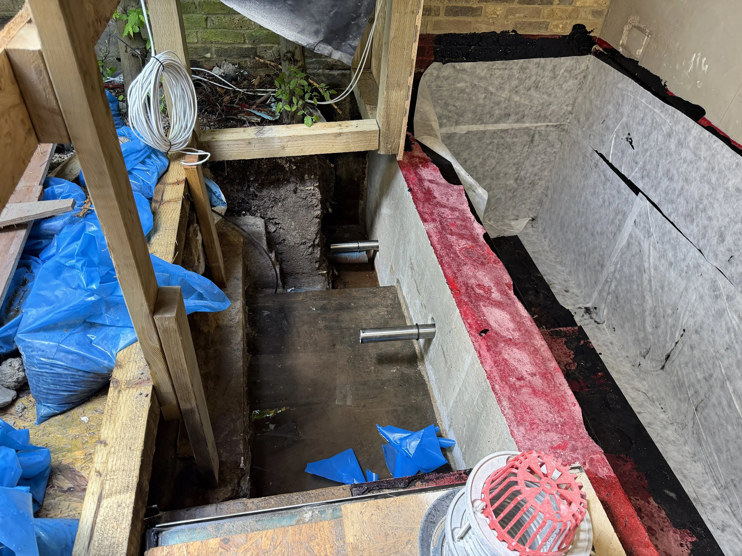 Construction site with wooden framing, exposed brick wall, and insulation. There are electrical wires, blue plastic sheets, and tools on the floor, with a person wearing orange shoes standing nearby.