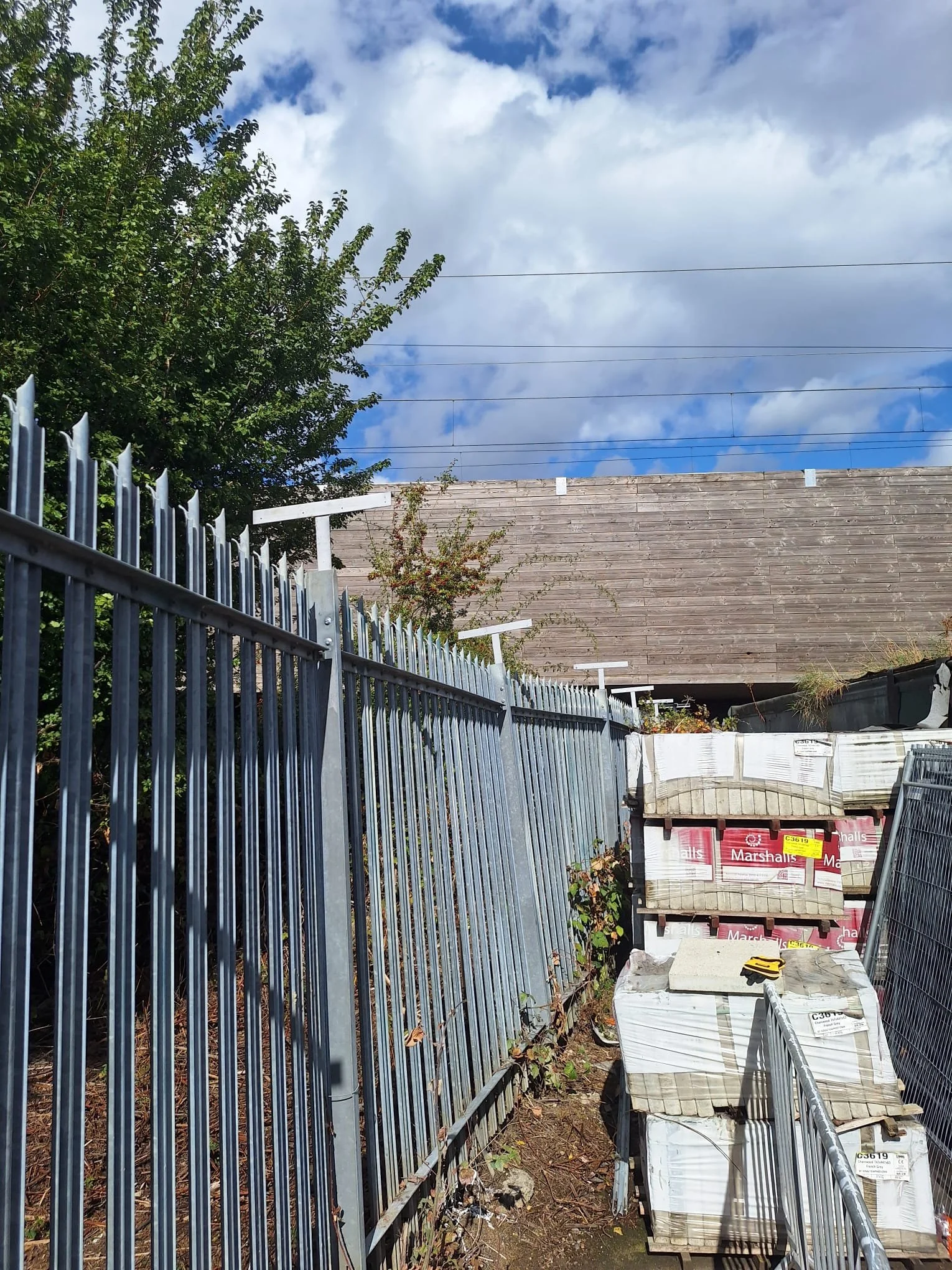 A construction site with a metal fence, a wooden wall, and a pile of marshmallow bags. The sky is partly cloudy with blue patches, and there is a tree on the left side.