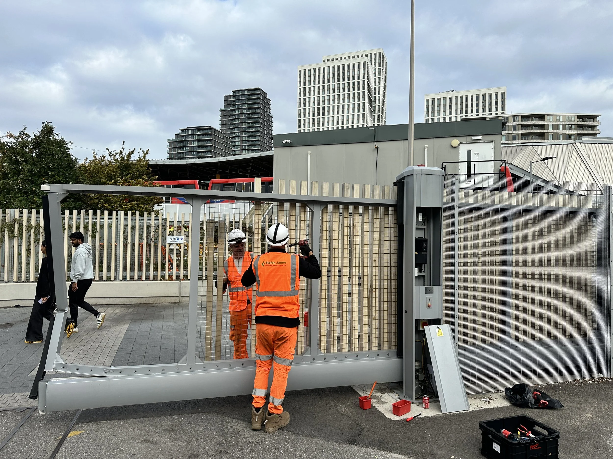 Construction workers installing a security gate outdoors in an urban area with tall buildings and cloudy sky in the background.