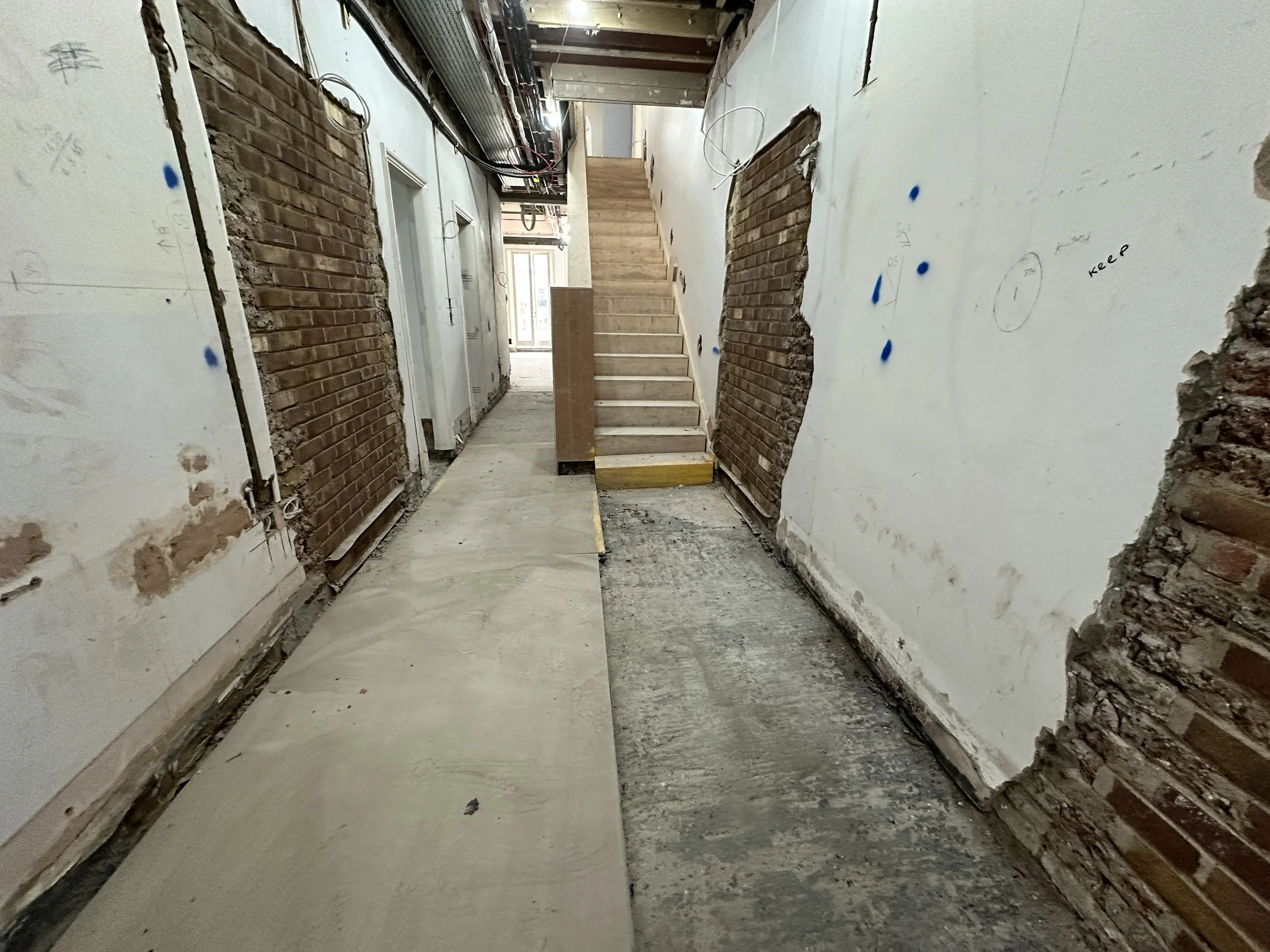 Construction site hallway with exposed brick walls on both sides, some drywall, and visible electrical wiring, leading to a staircase in the background.
