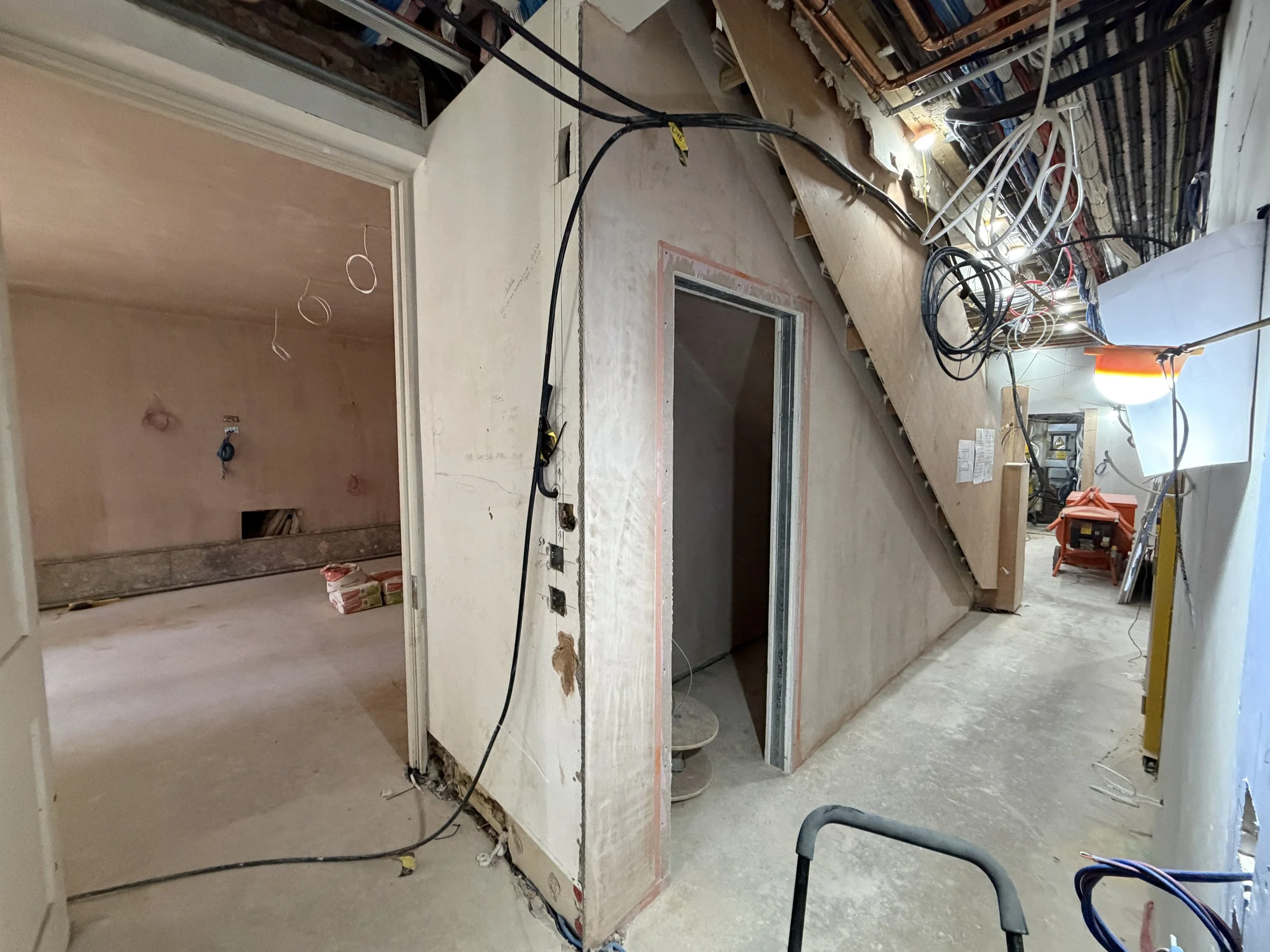 This image shows an under-construction or renovation interior with electrical wiring and building materials. There is a partially built wall with exposed frames and electrical outlets, and construction tools and supplies are scattered around.