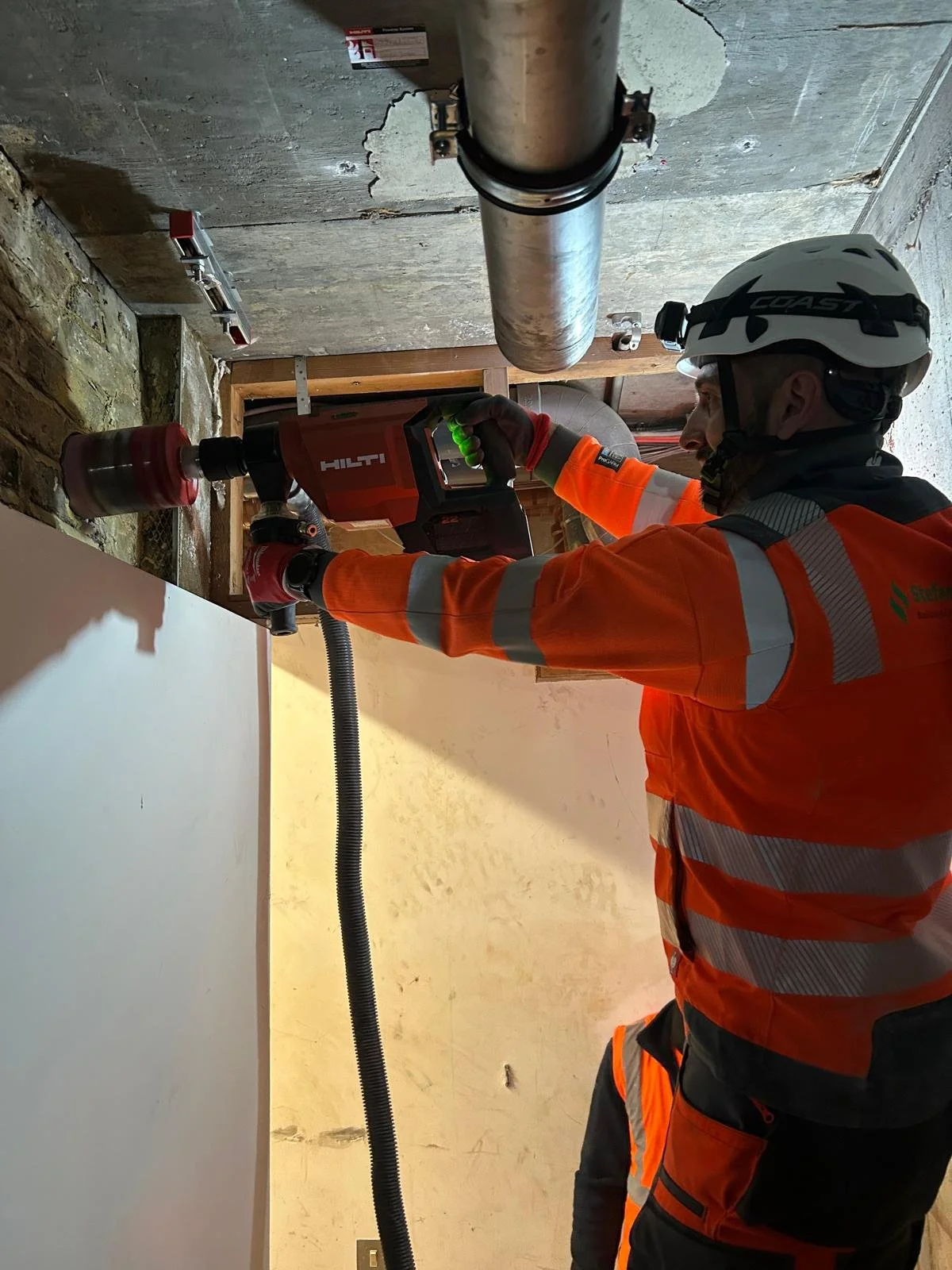A construction worker wearing a safety helmet and orange high-visibility jacket uses a power drill to drill into a wall in a building under construction.